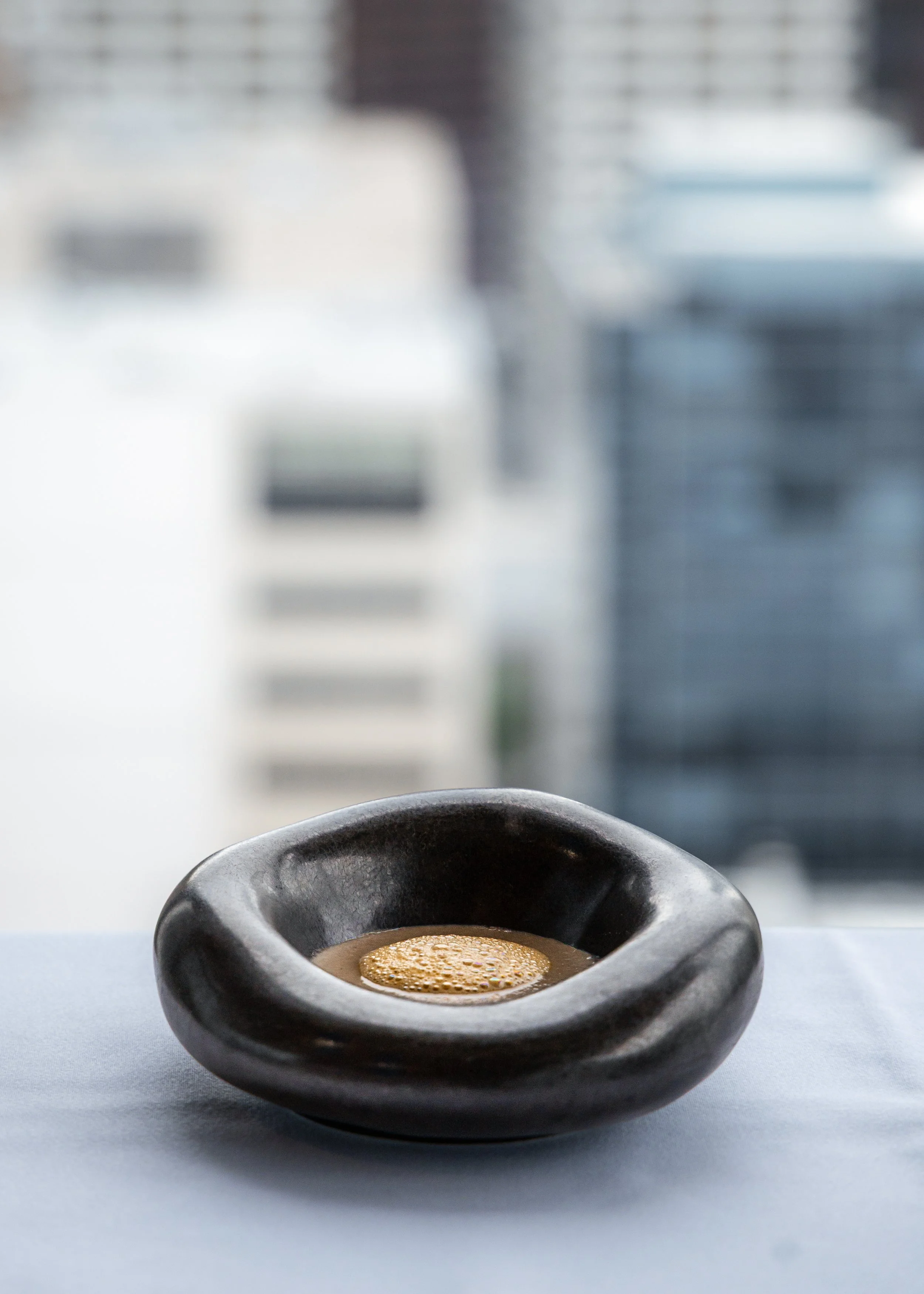 A small, black ceramic bowl filled with frothy, brown soup, placed on a light blue surface with a blurred modern interior background.