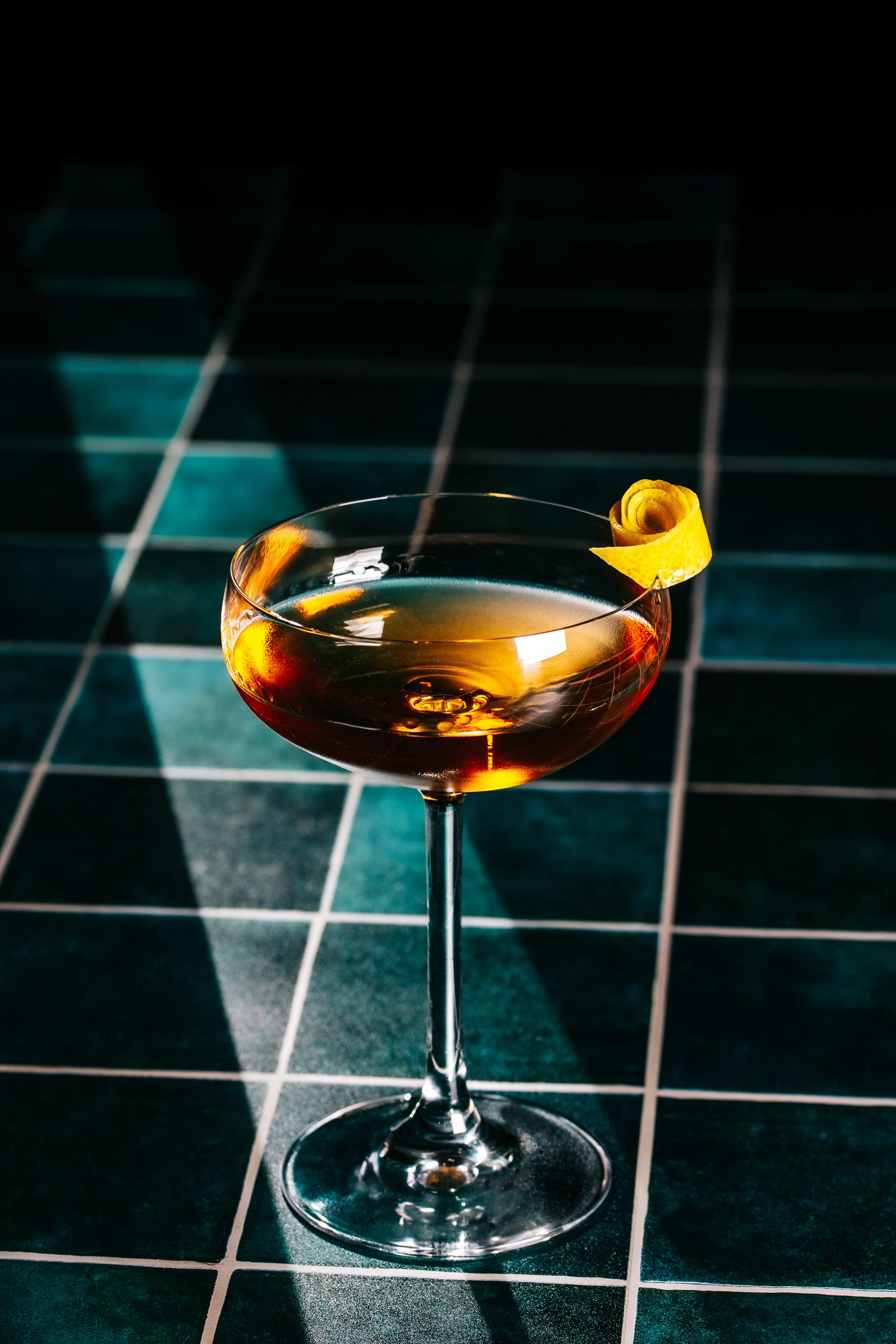 A cocktail glass filled with a dark amber drink, garnished with a yellow lemon twist, set on a dark tiled surface with dramatic lighting and shadows.