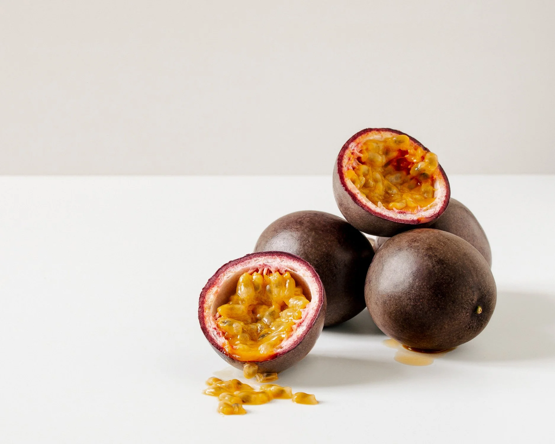 A group of whole passion fruits with one cut open, revealing bright yellow pulp and seeds inside, arranged on a white surface against a plain light background.