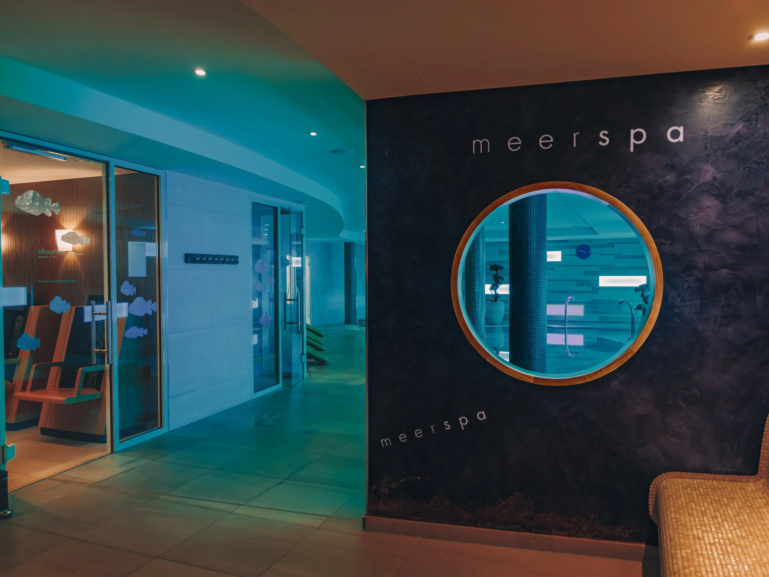 Indoor spa area with a dark textured wall displaying the name 'meerspa', a large circular mirror, and a seating area with coral-colored chairs.