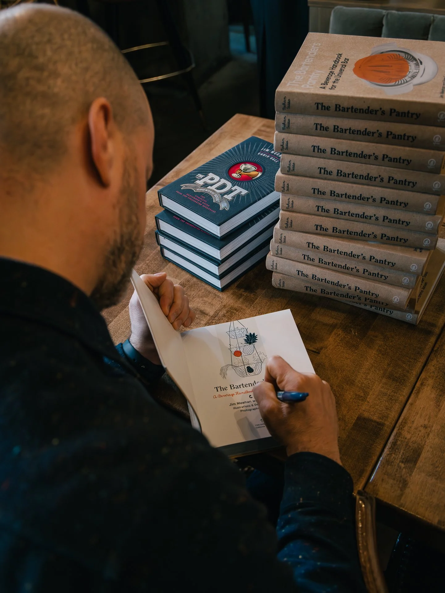 A person reading a book titled 'The Bartender's Pantry' in a restaurant or cafe setting, with multiple copies of the same book stacked on the table. The person is holding a pen and appears to be reviewing or signing the book.