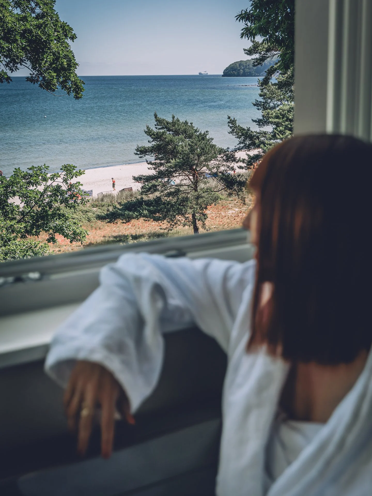 A woman with brown hair relaxing by a window, looking out at a beach with trees, sand, and water, with a person walking on the beach and a ship in the distance.