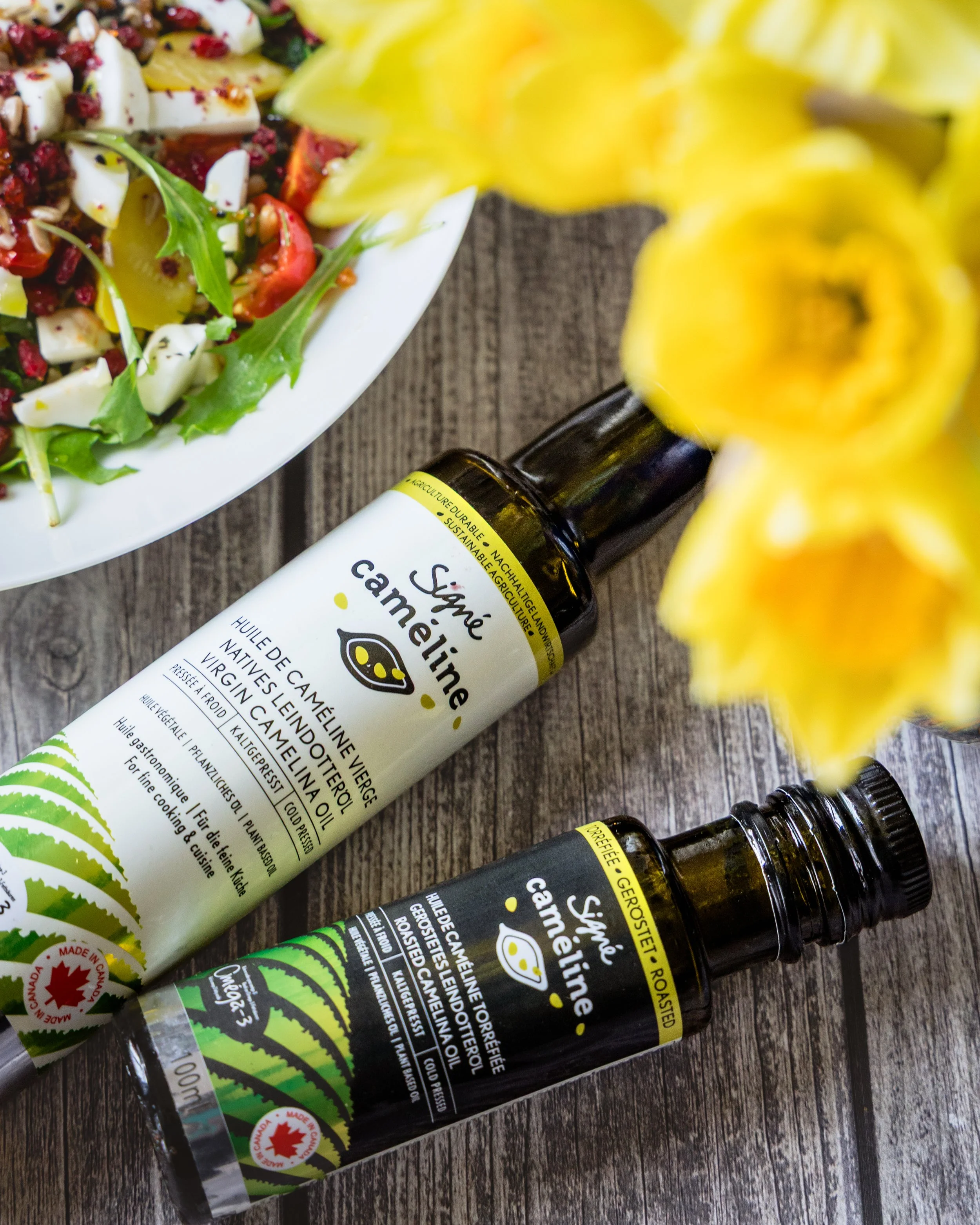Close-up of two bottles of Camelina oil and a salad with tomatoes, cheese, and arugula on a wooden surface.