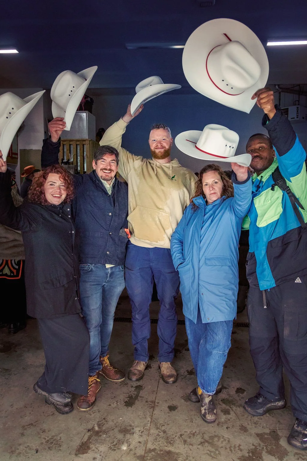 Group of five people celebrating indoors, holding cowboy hats in the air, smiling and raising their arms.