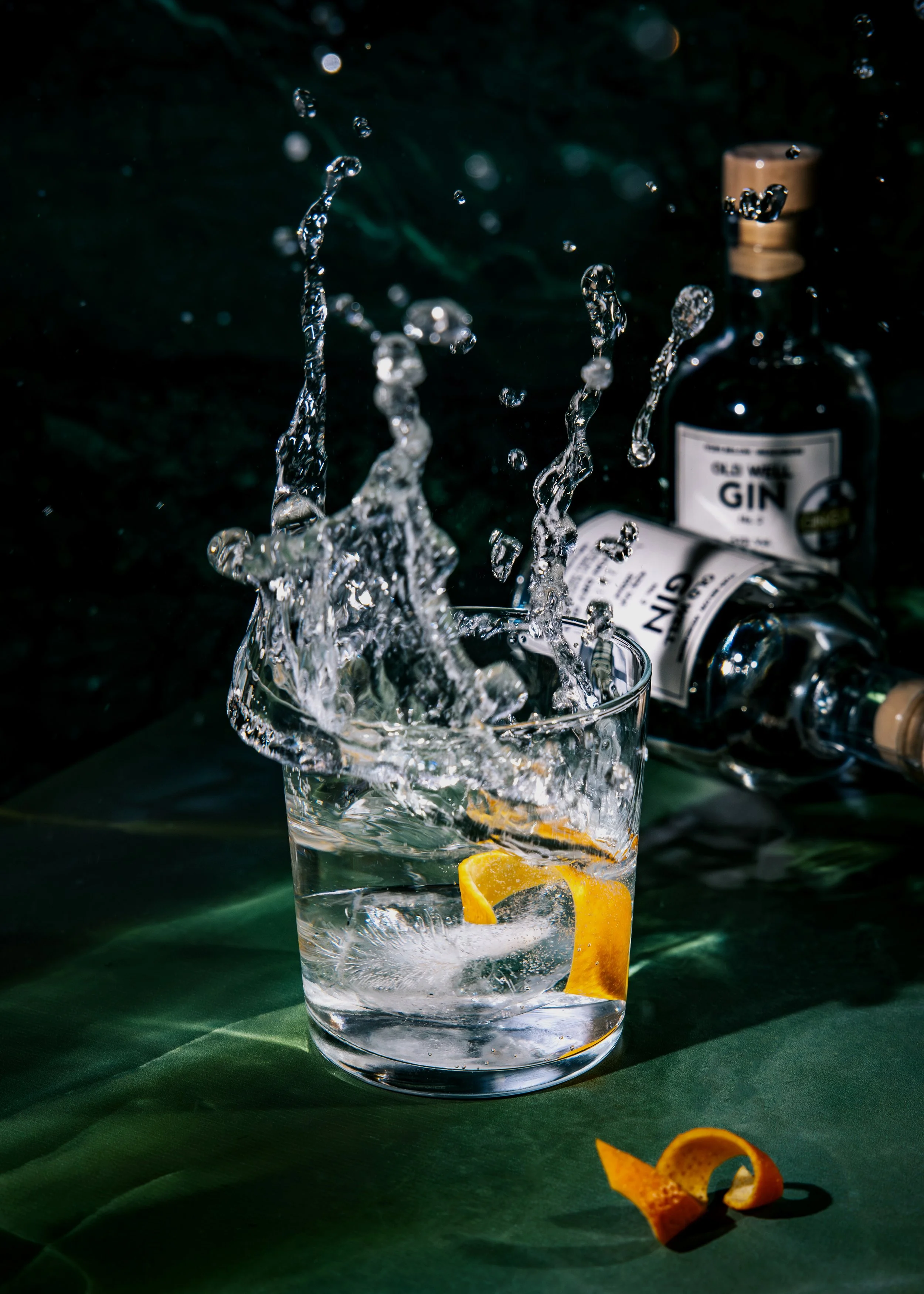 A glass of water with a lemon wedge inside, with water splashing out of the glass. In the background, there is a bottle of gin knocked over on a dark surface, with lemon peel on the table.
