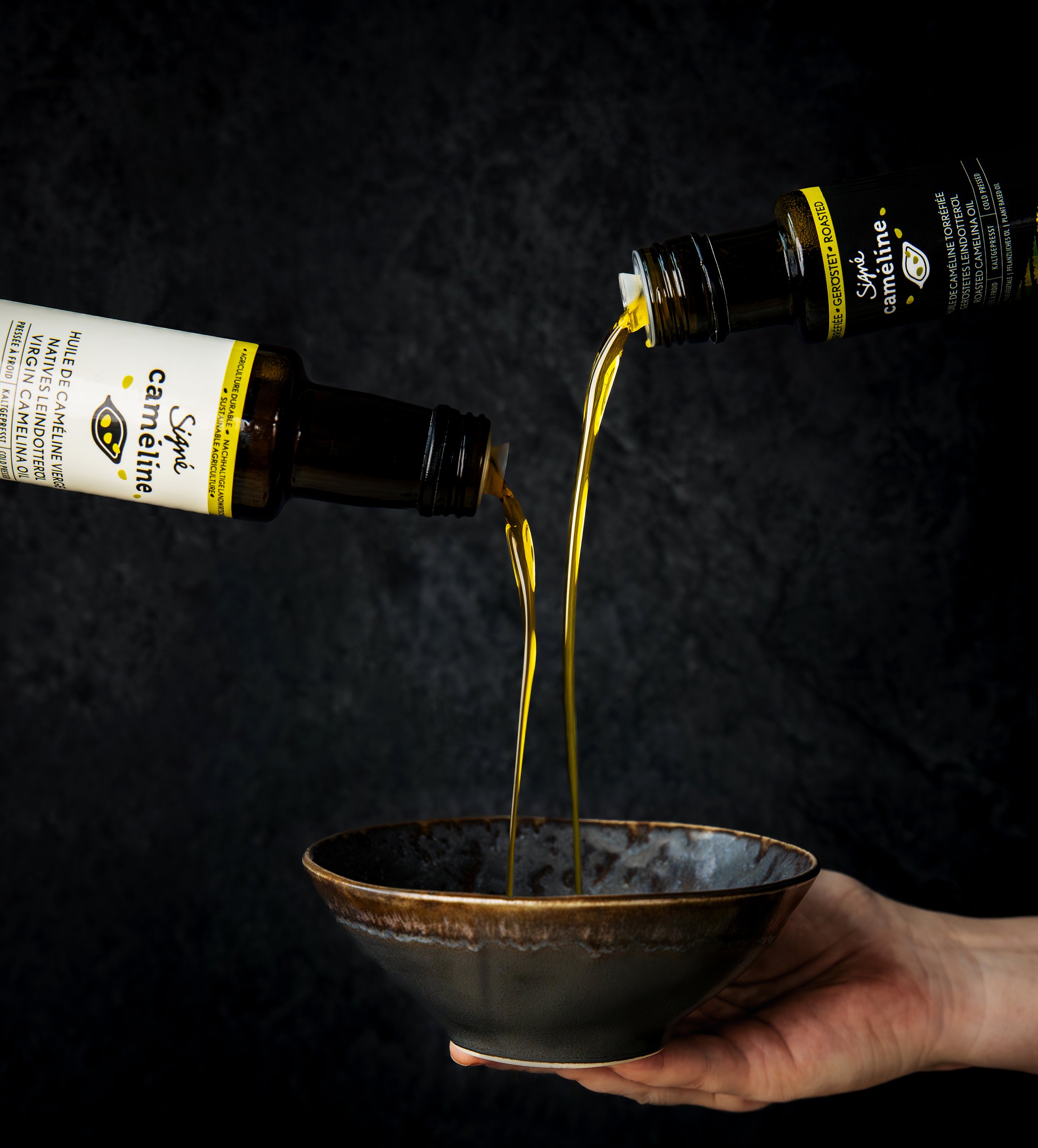 A hand holding a ceramic bowl with dark glossy glaze, while golden liquid is being poured into the bowl from a small dark bottle labeled 'SignéCamelline Roasted' oil.