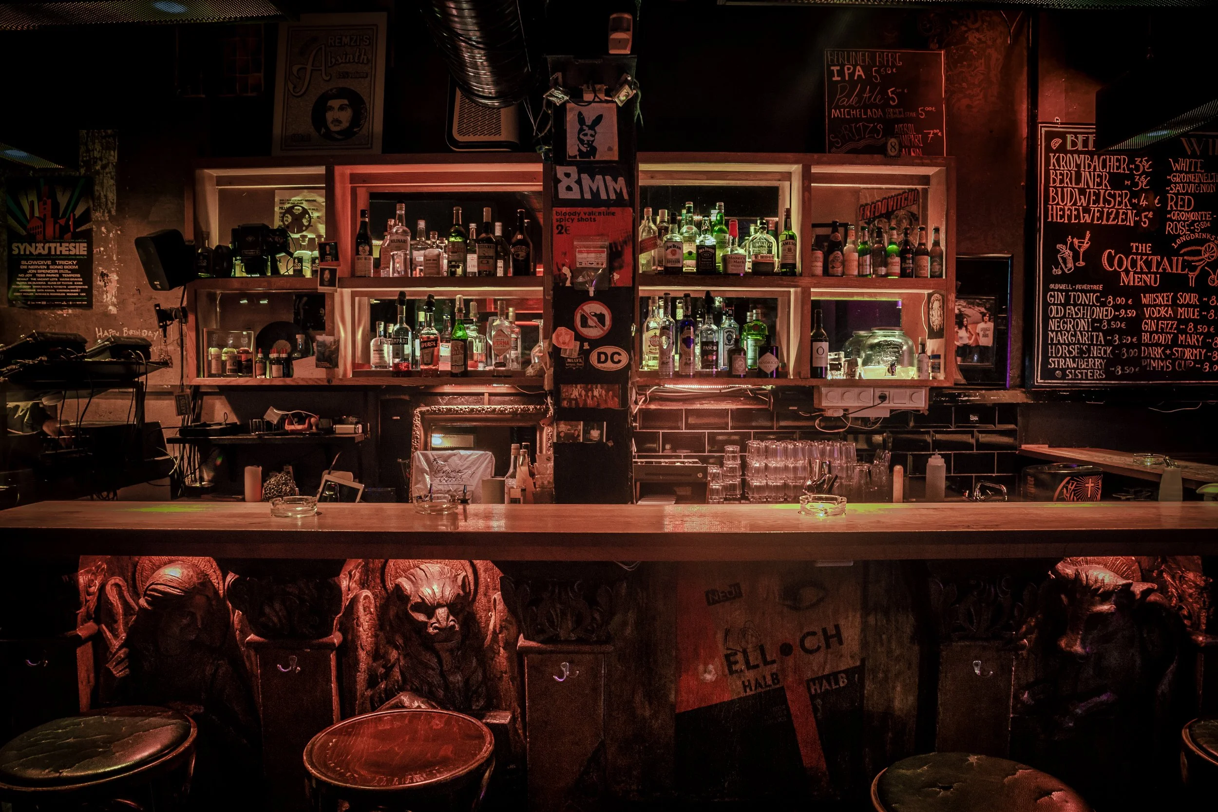 A bar with shelves of various liquor bottles, illuminated with dim lighting, in a cozy, dark ambiance. There are black bar stools in the foreground and chalkboard menus on the walls.