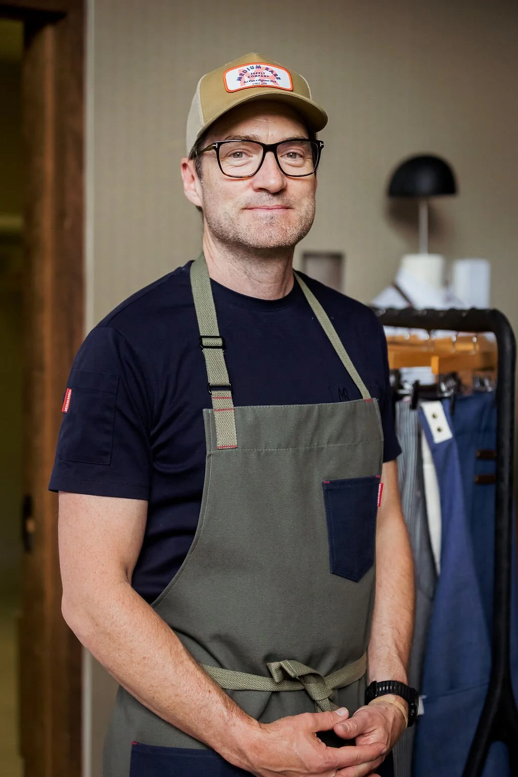 A man wearing glasses, a tan cap, a dark T-shirt, and a green apron standing indoors with a clothing rack in the background.
