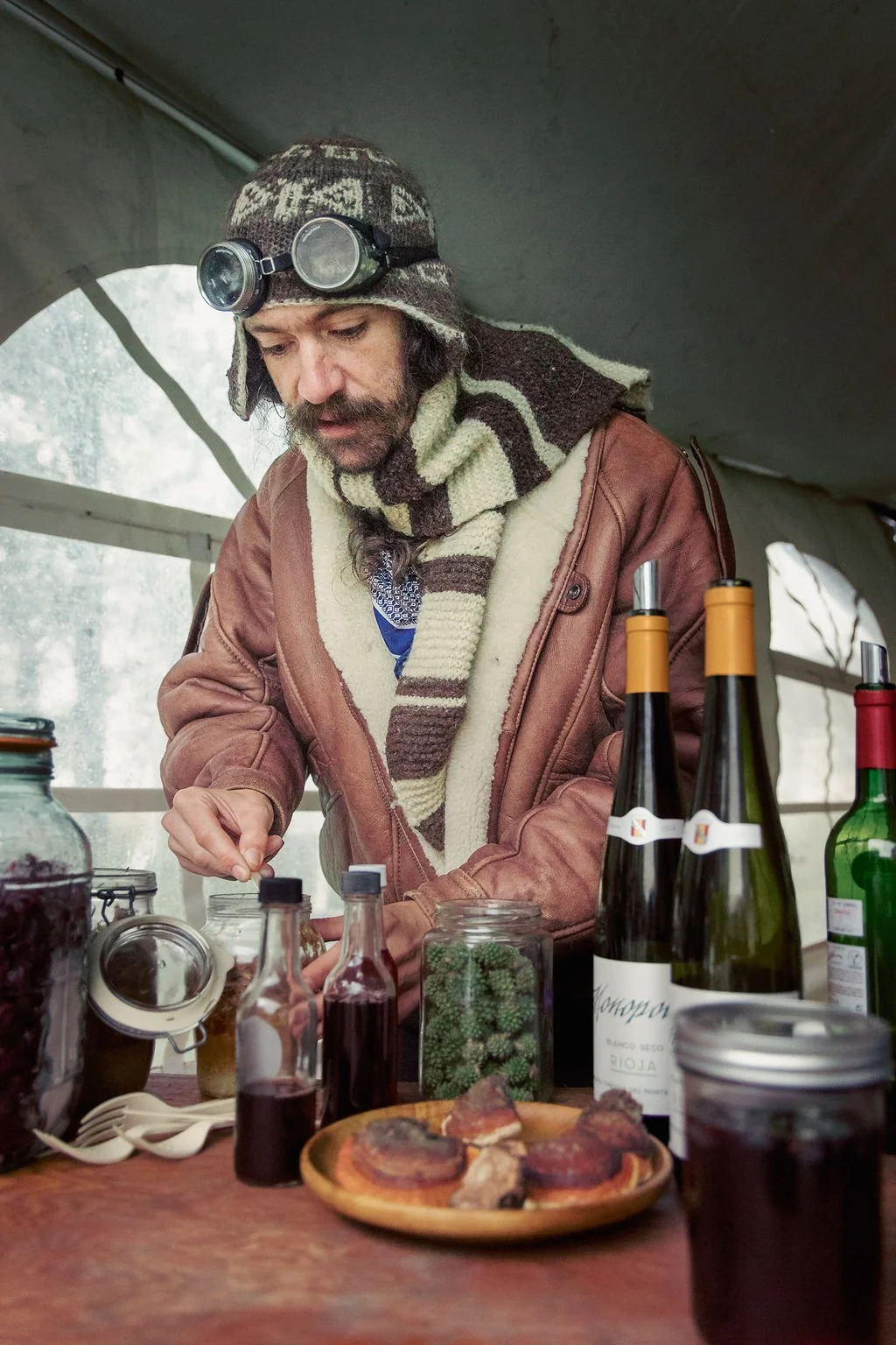 A man wearing a brown shearling jacket, striped scarf, and wool hat with goggles inspects bottles and jars on a table inside a tent.