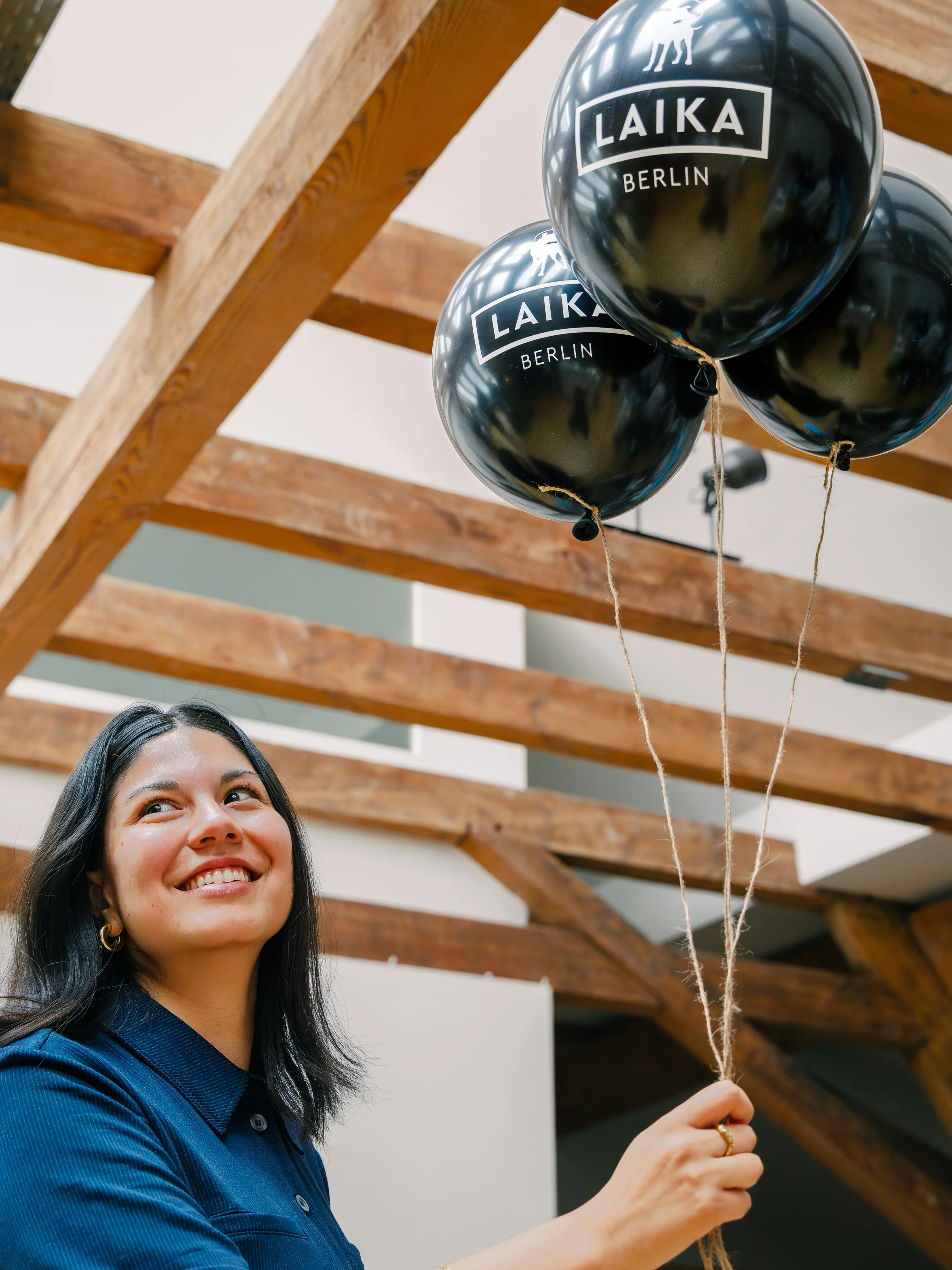 A woman smiling and holding black balloons with the text 'LAIKA BERLIN' printed on them, standing indoors under a wooden ceiling.