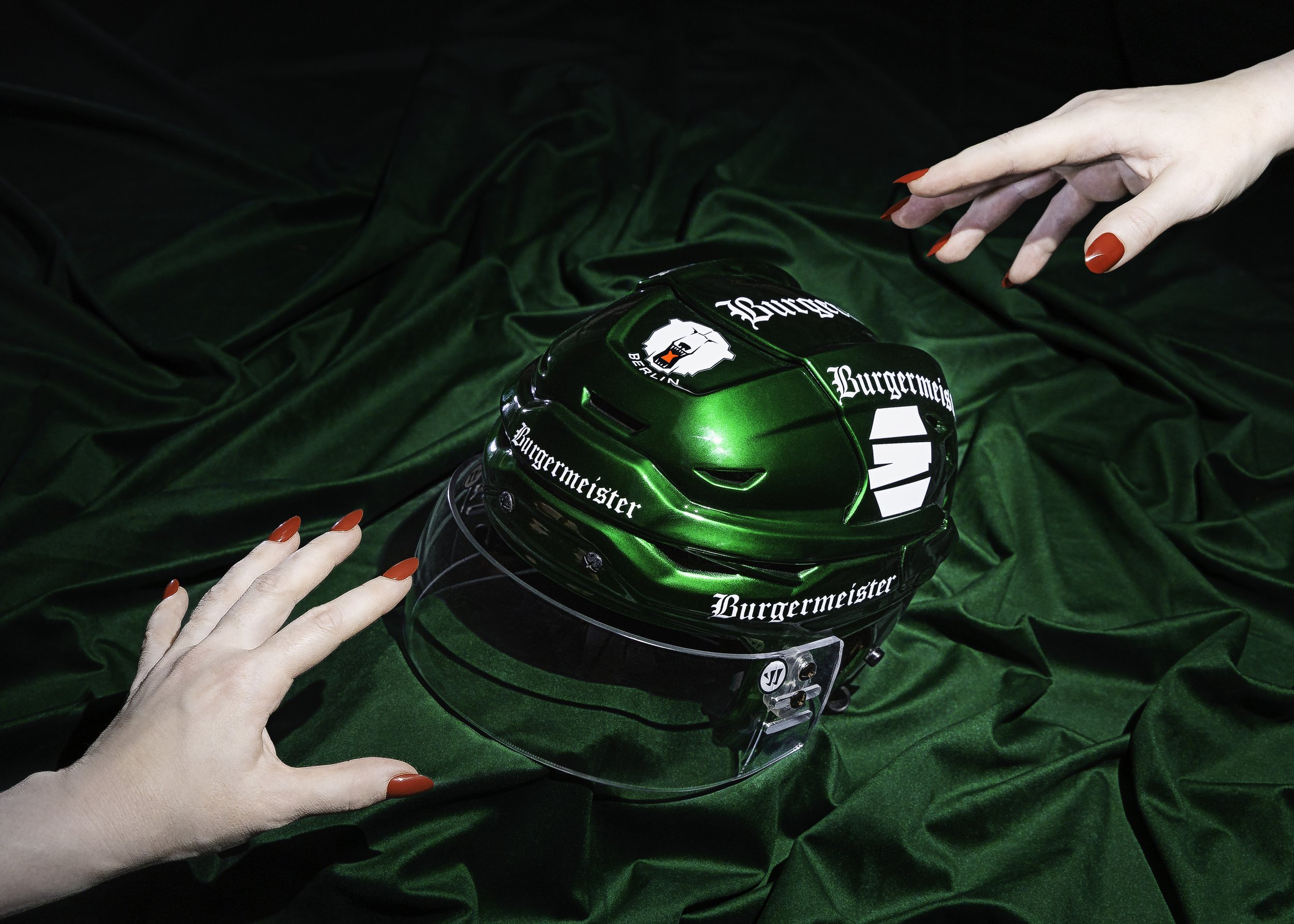 A woman with red nails reaches towards a green hockey helmet with the brand 'Burgermeister' and a logo featuring a polar bear's head and the word 'Berlin' on a dark green fabric background.