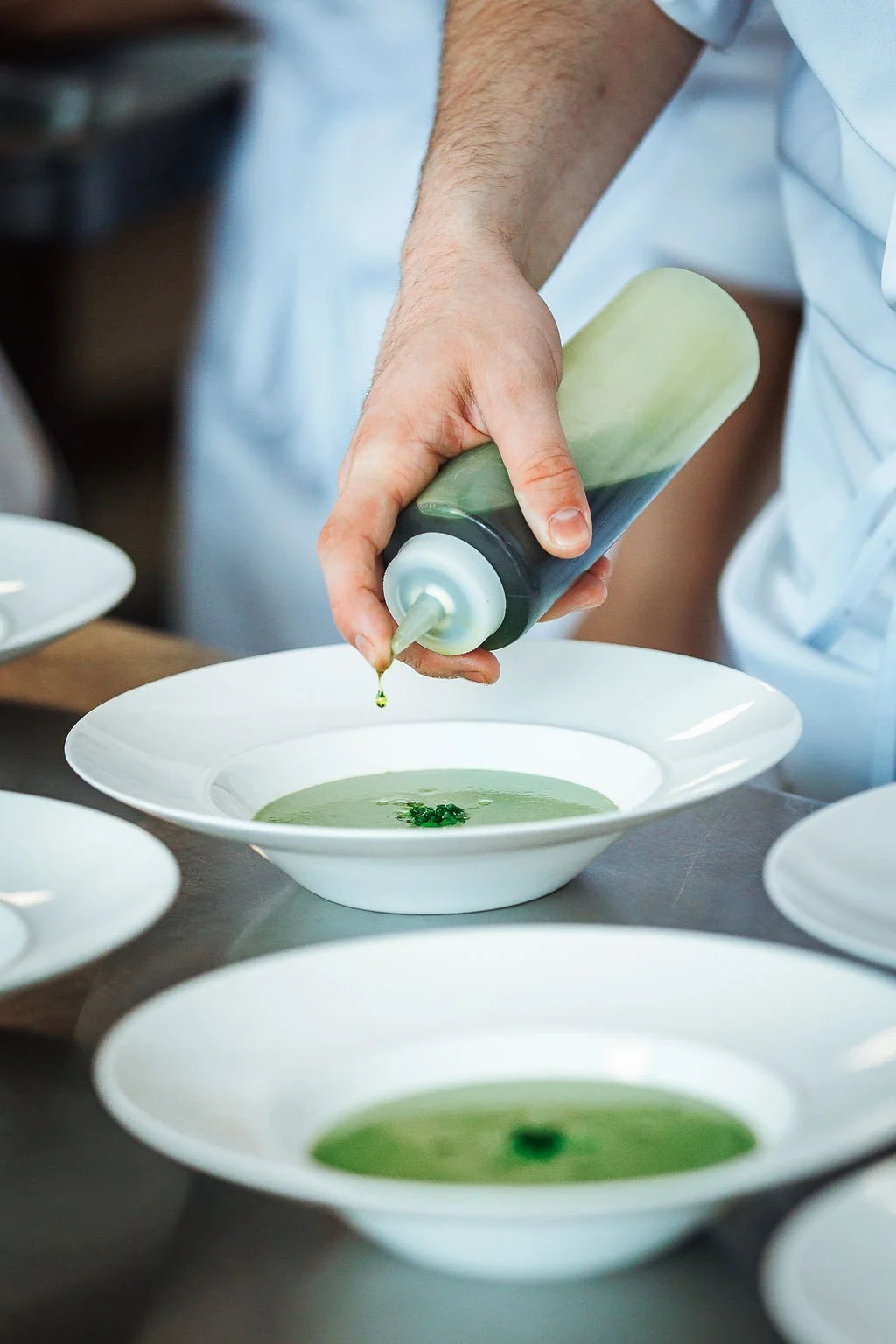 Chef pouring green sauce onto white soup bowls in a kitchen.