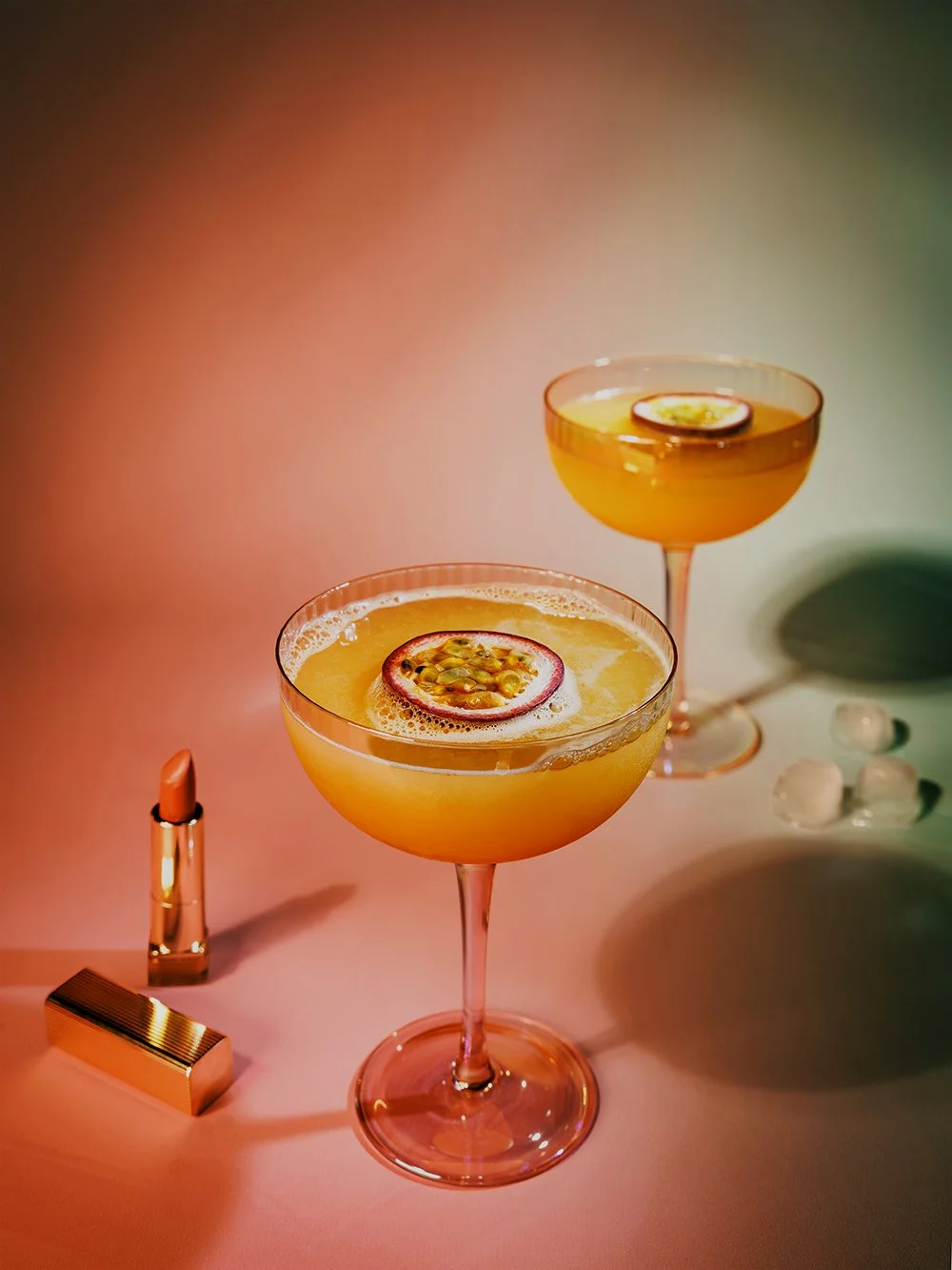 Two yellow cocktails garnished with passion fruit slices, surrounded by lipstick, a gold-colored makeup brush, and ice cubes on a colorful backdrop.