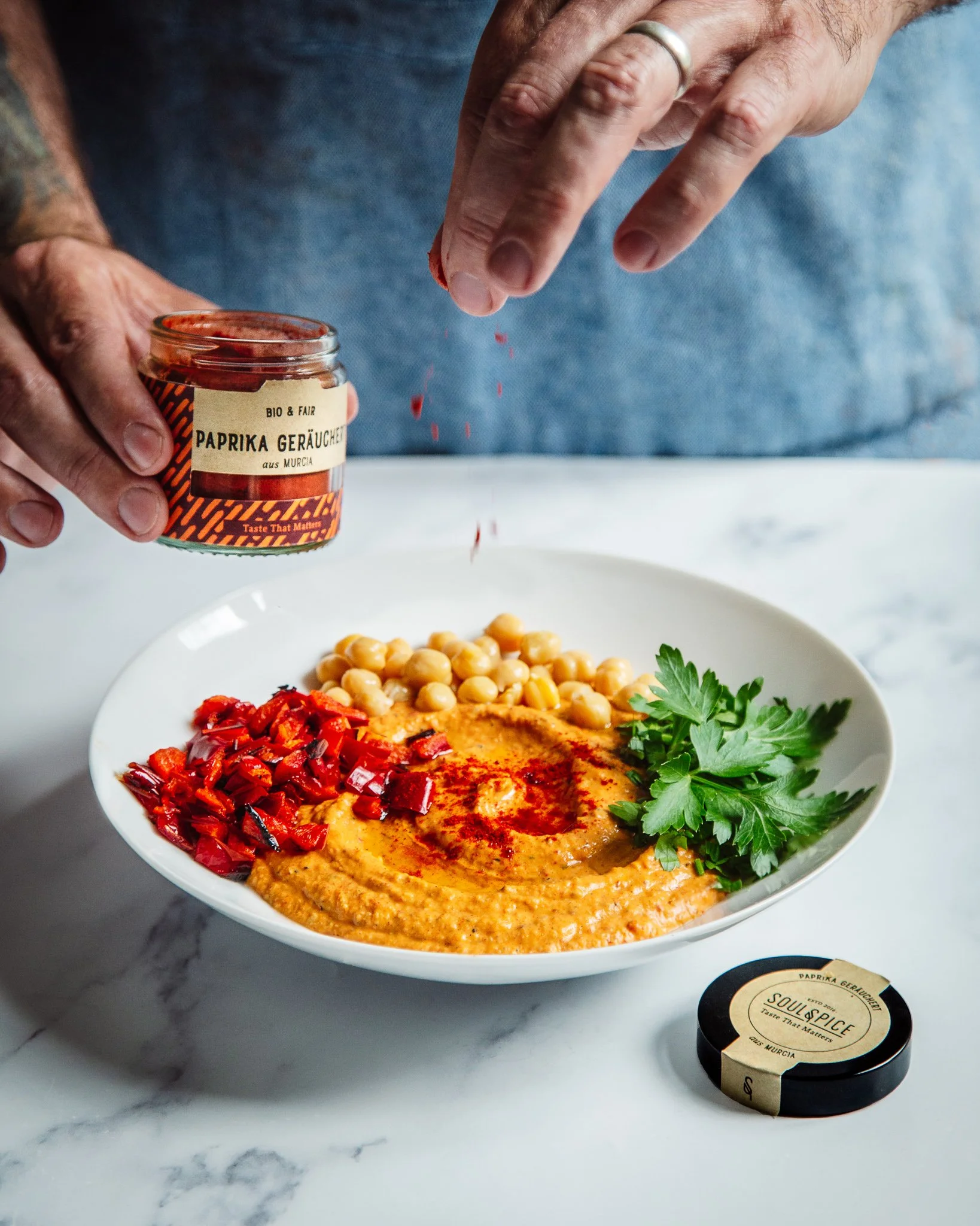 A white bowl containing hummus, chopped red peppers, and fresh parsley, on a marble countertop. A hand is sprinkling paprika from a small jar labeled "Paprika Geräuchert" onto the hummus.