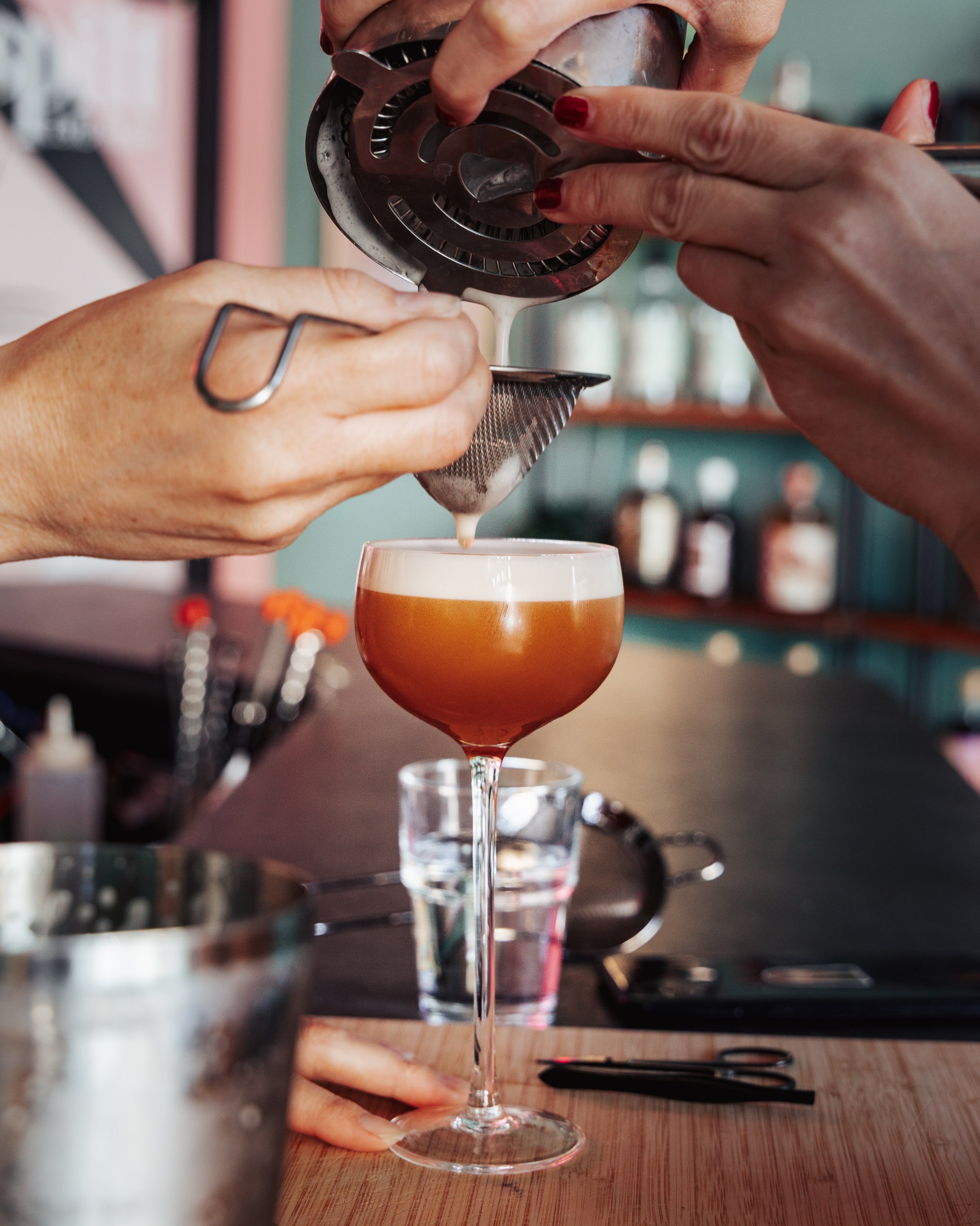 A person pouring a light cream-colored liquid, likely a cocktail or beverage, into a glass. The person's hands are visible, with red nail polish. The image is focused on the act of pouring, with a bar or kitchen background.
