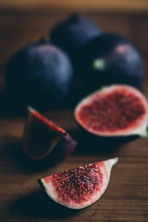 Close-up of fresh figs, with one sliced open showing red flesh and seeds, on a wooden surface with whole purple figs in the background.
