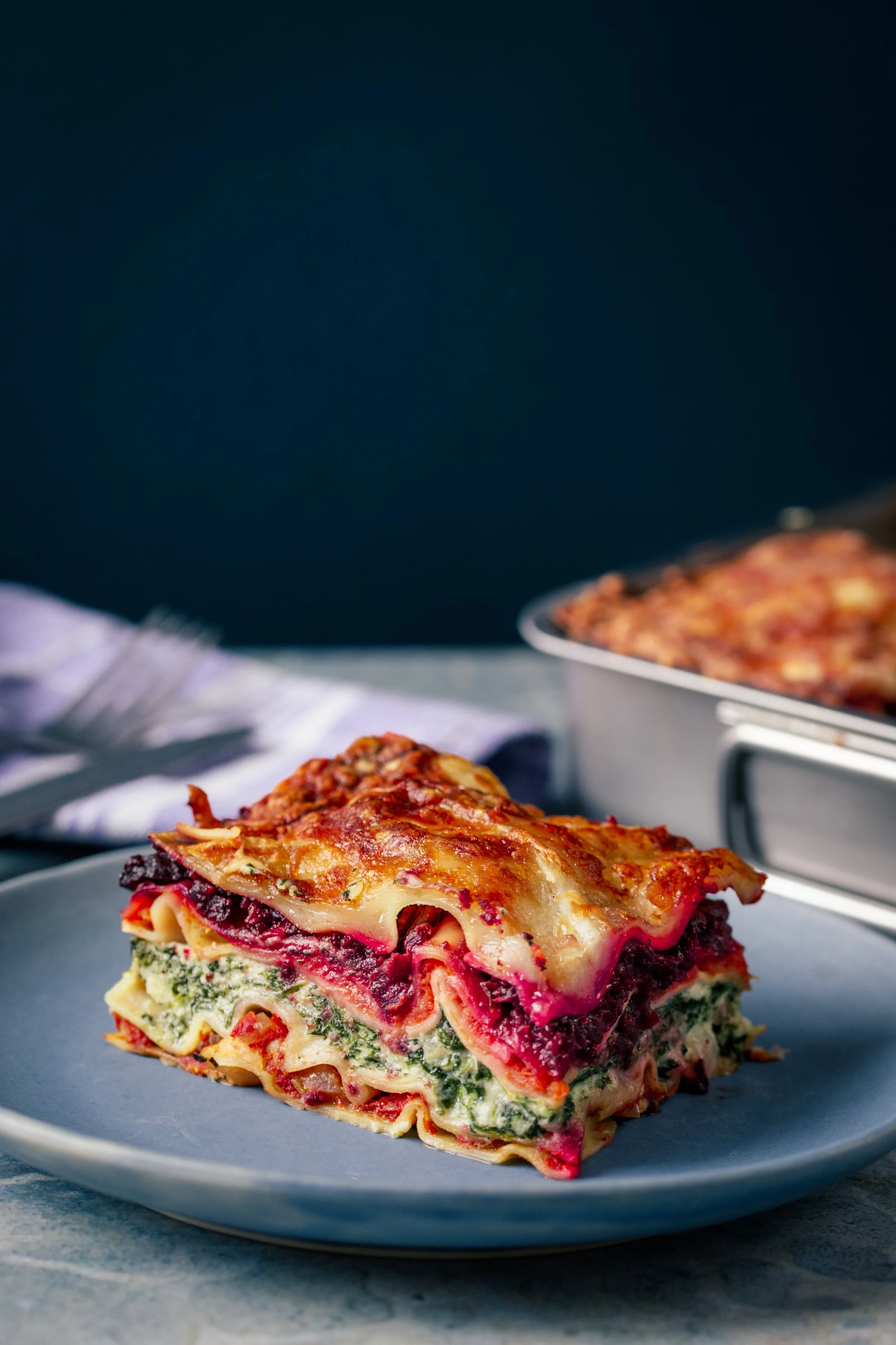 A serving of vegetable lasagna on a gray plate with layers of pasta, spinach, tomato sauce, and cheese, topped with melted cheese. In the background, more slices of lasagna are on a baking dish.