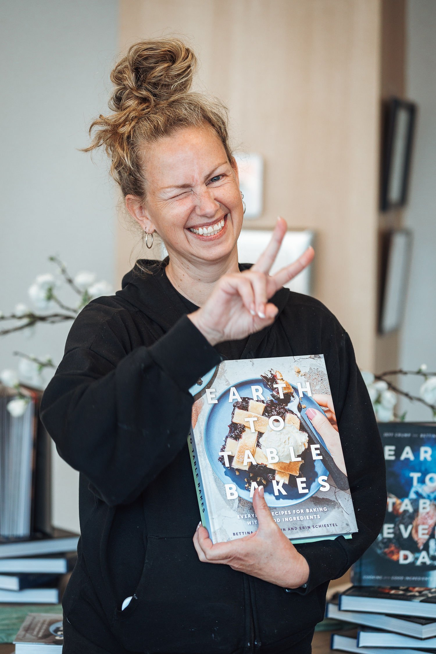 Woman with curly hair in a messy bun holding a cookbook titled "Earth to Table Bakes" and making a peace sign gesture with her right hand.