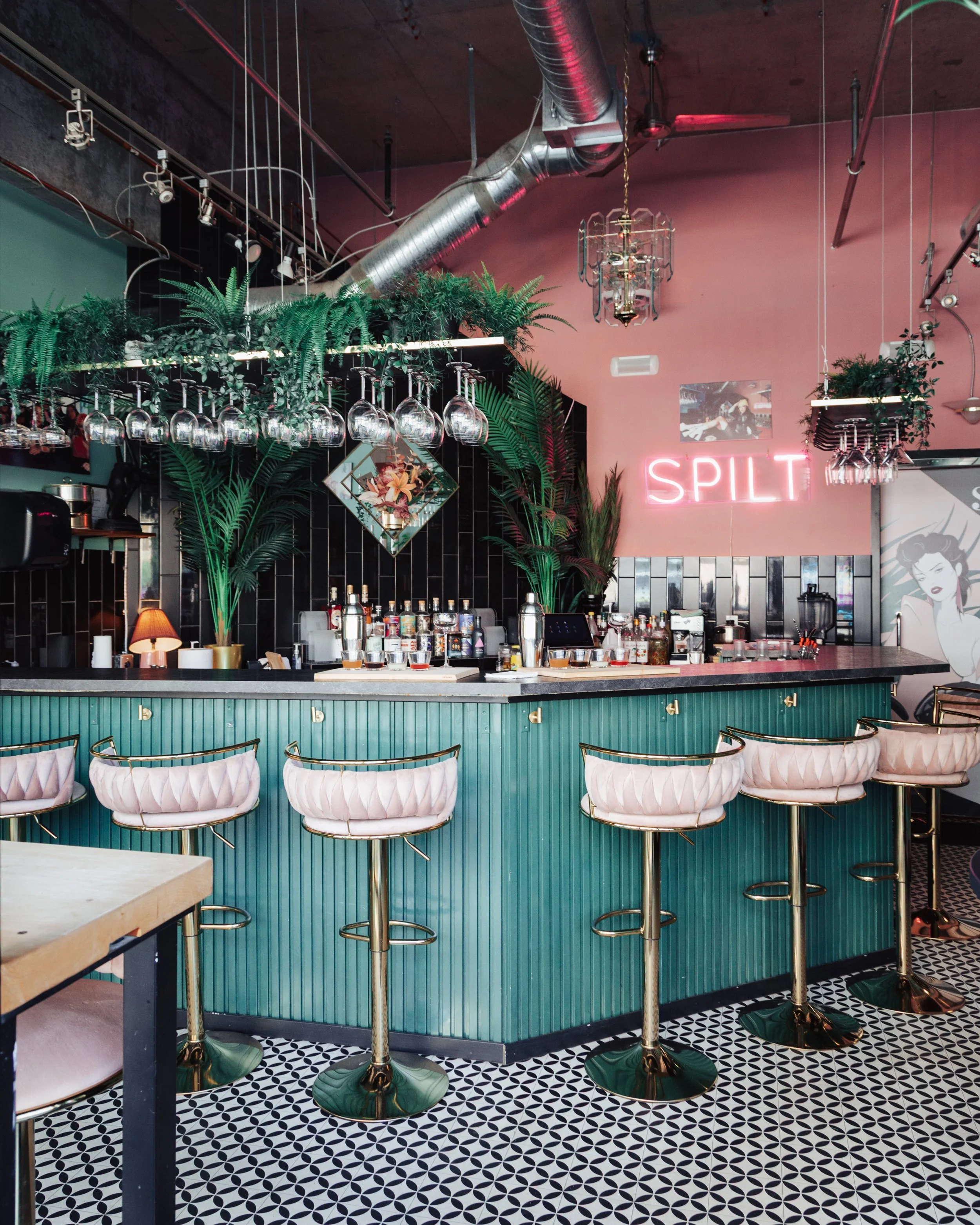 Interior of a stylish bar with a mint green counter, white and pink cushioned bar stools, hanging wine glasses, and lush green plants, with a pink neon sign that says 'SPLIT' on the wall.