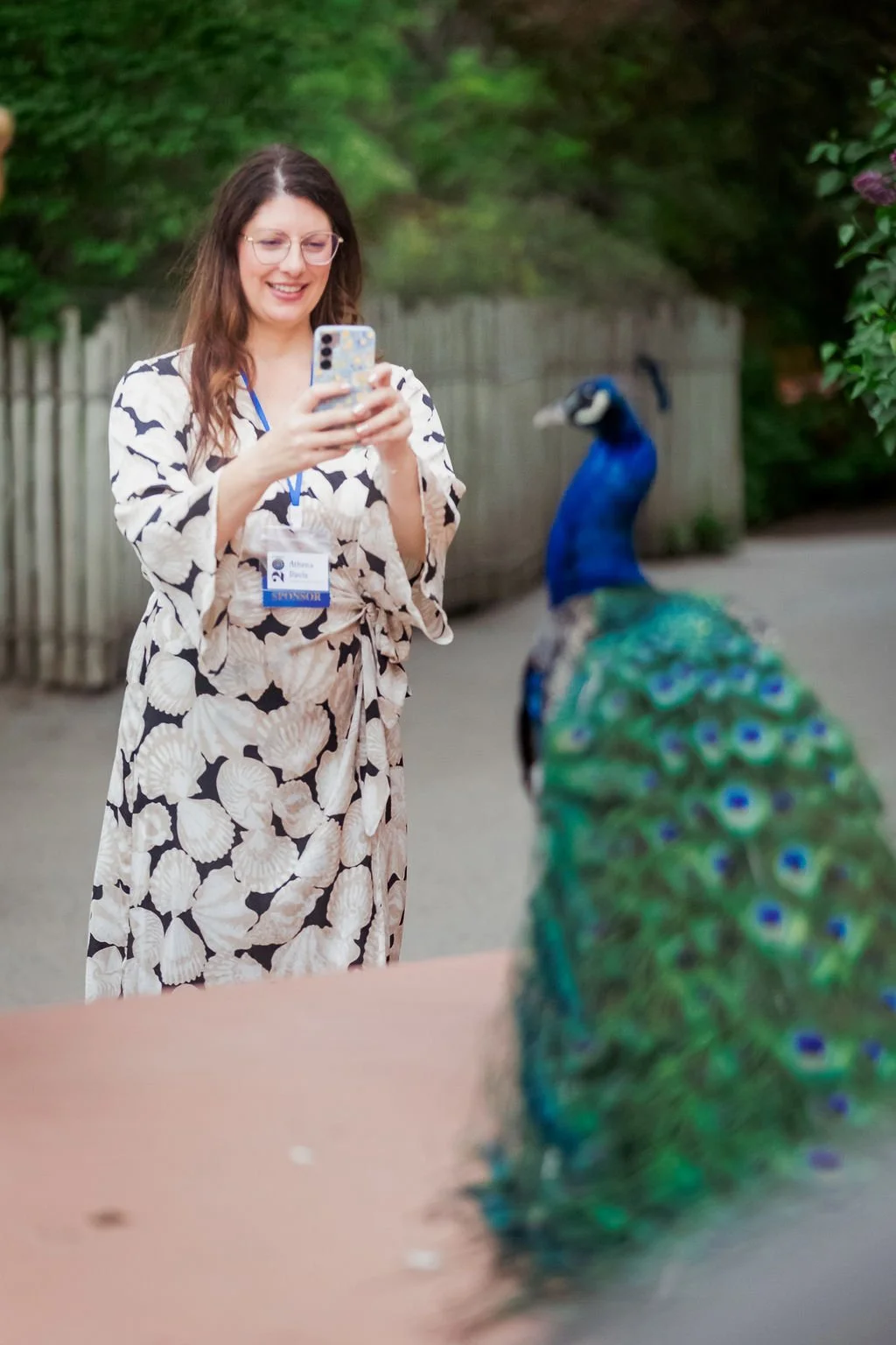 A woman with glasses and a seashell patterned dress is smiling and looking at her phone while standing near a peacock.
