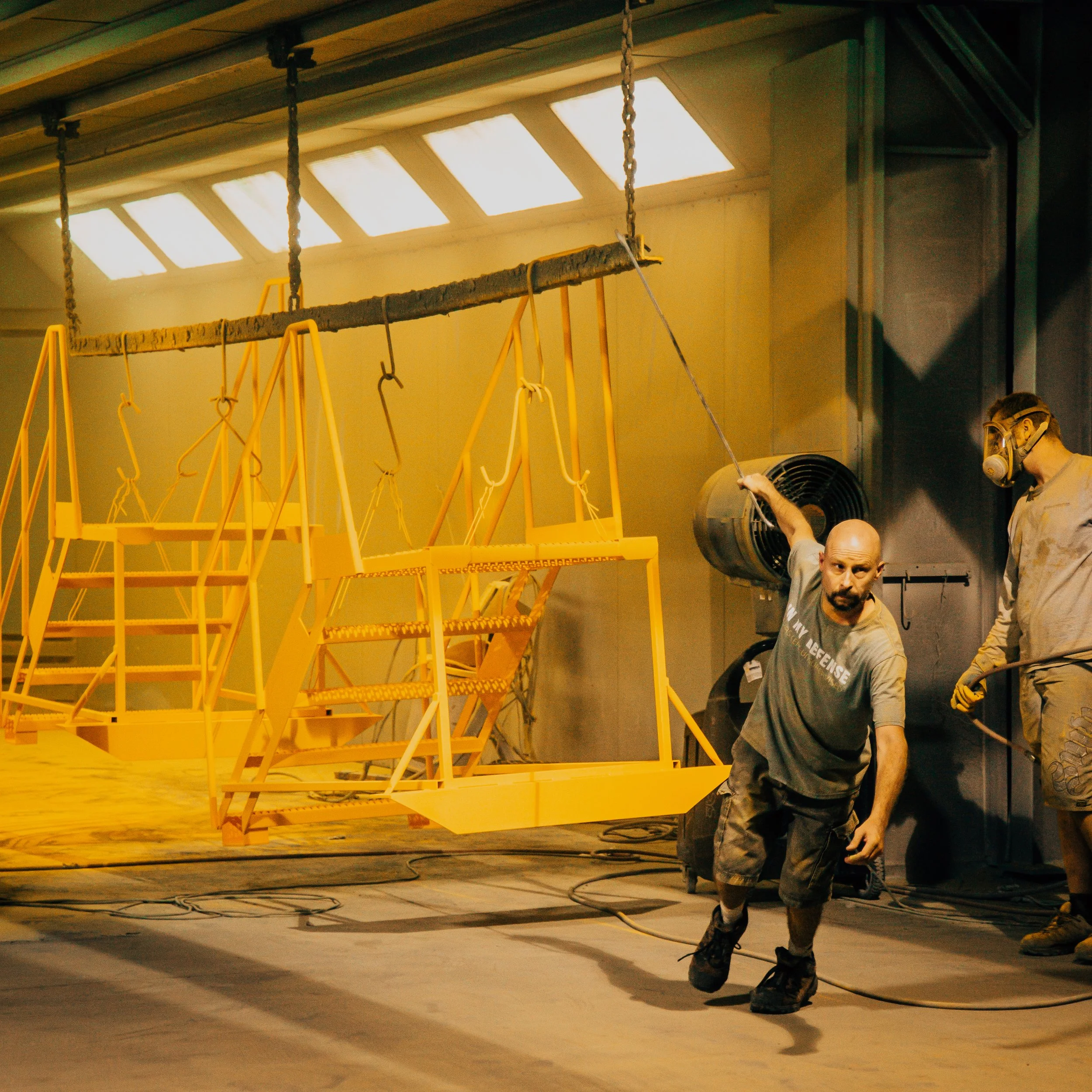 A construction worker wearing safety gear, including ear protection and gloves, stands in front of a yellow scaffolding structure inside a building. The ceiling has large skylights allowing natural light to illuminate the workspace.