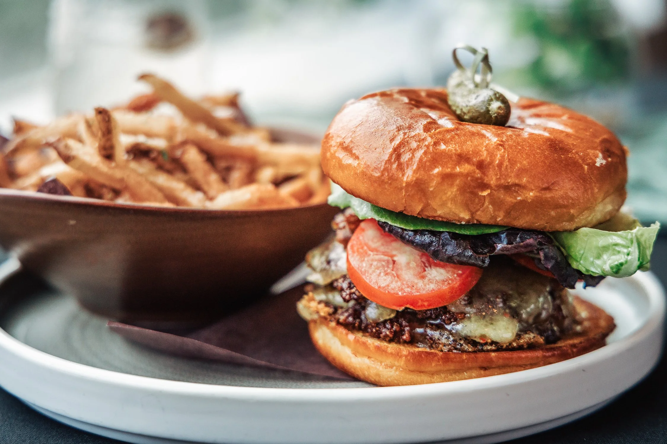 A close-up of a gourmet burger with lettuce, tomato, and grilled patty, topped with a brioche bun and a pickle on top, served on a white plate with a side of pasta salad in the background.