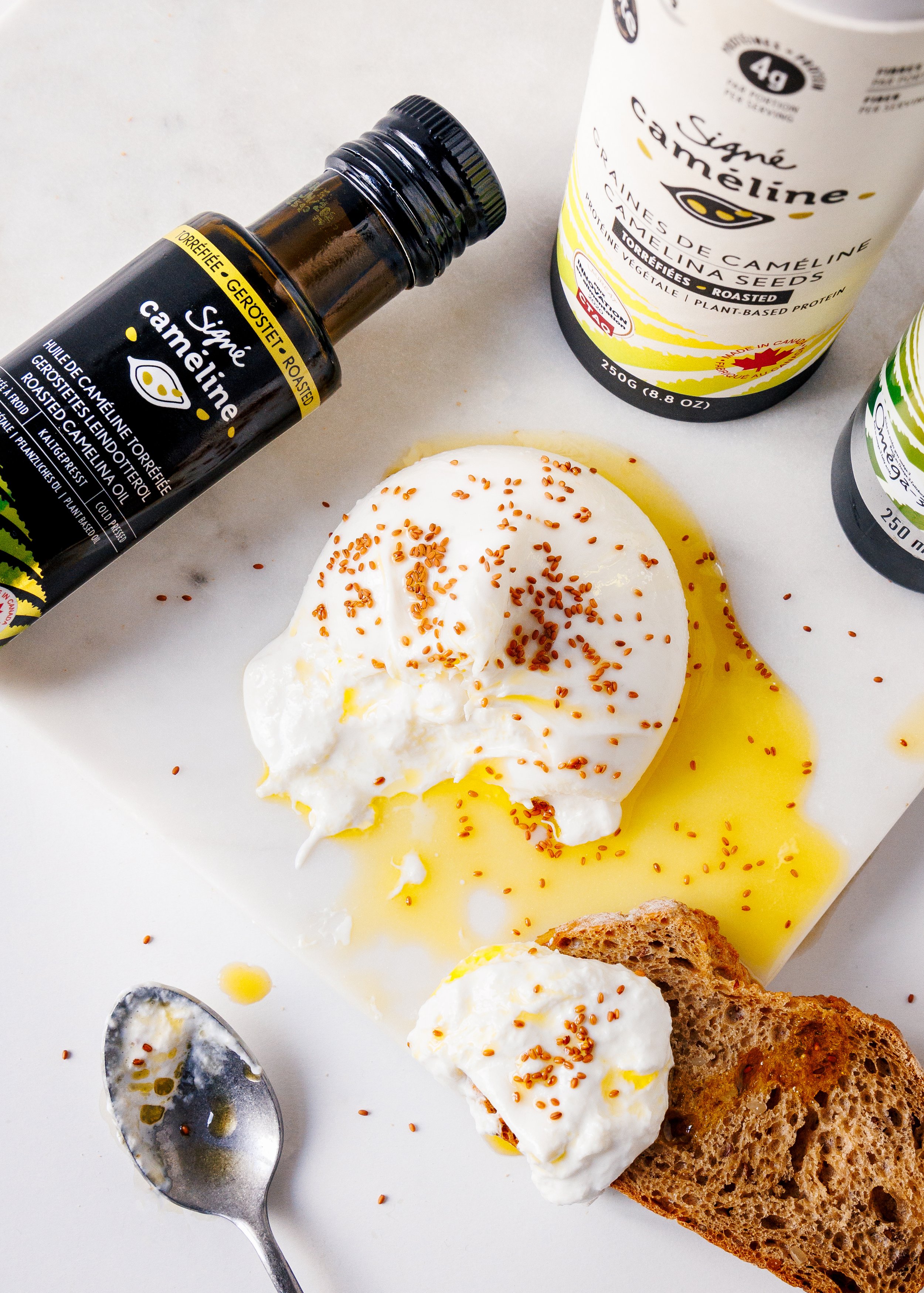 A breakfast scene with poached eggs topped with sesame seeds, a slice of whole grain bread, and a bottle of Signé Cameline roasted camelina oil. There is a spoon and an additional piece of bread partially visible.