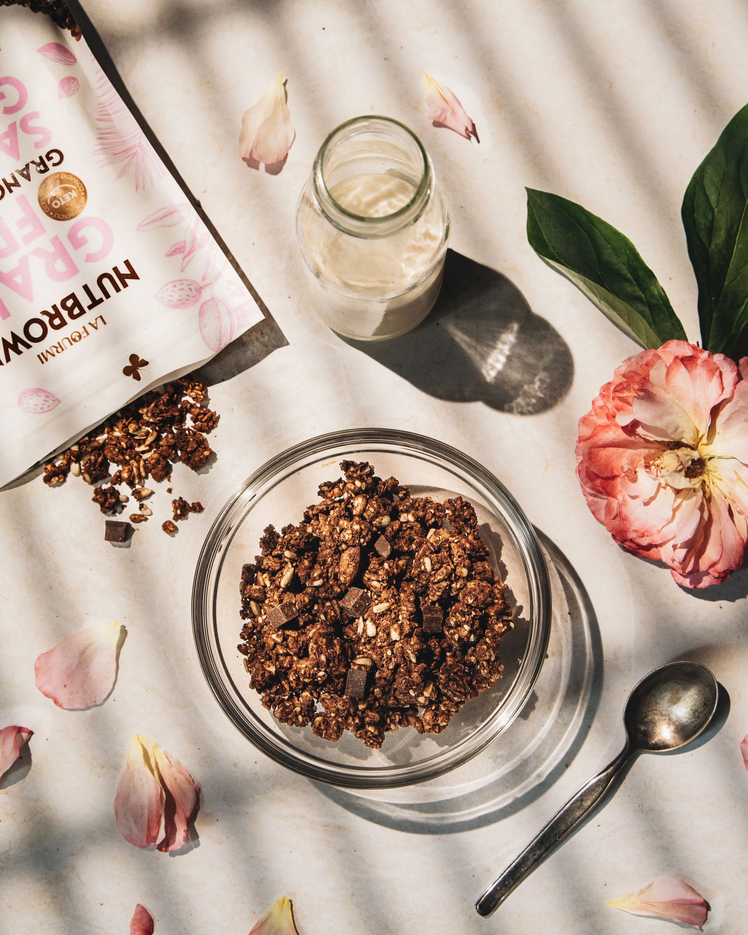 A glass bowl filled with chocolate granola on a white surface, surrounded by scattered granola pieces, pink and white flower petals, a silver spoon, and a partially visible bag of granola labeled 'Autumn Grain' with pink and brown lettering and pink 