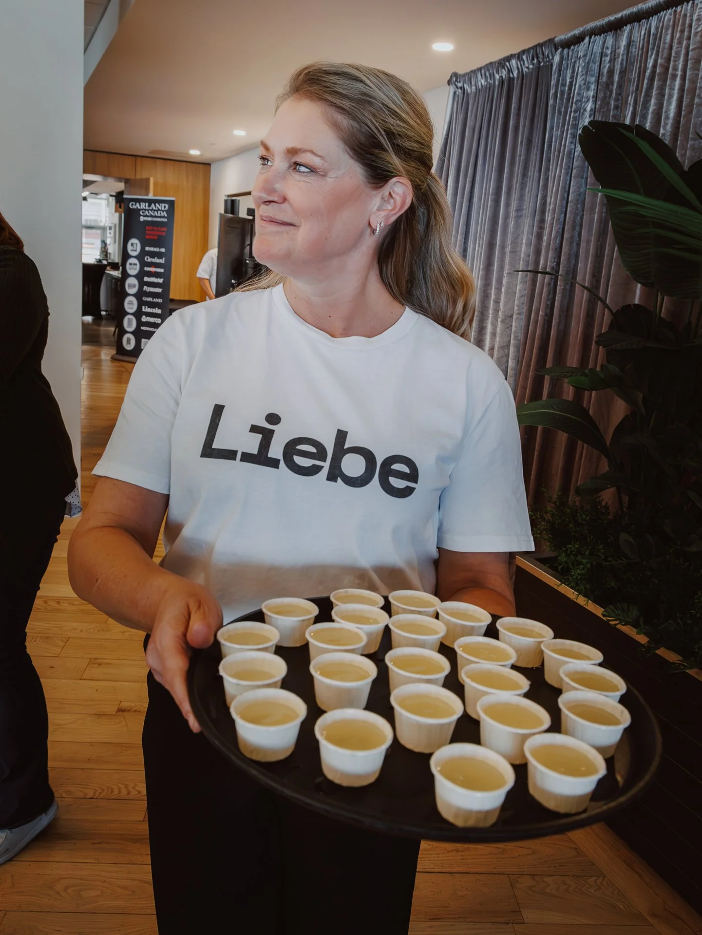A woman wearing a white shirt with the word 'Liebe' is holding a tray of small cups filled with a light-colored liquid, possibly soup or dessert, at an indoor event.