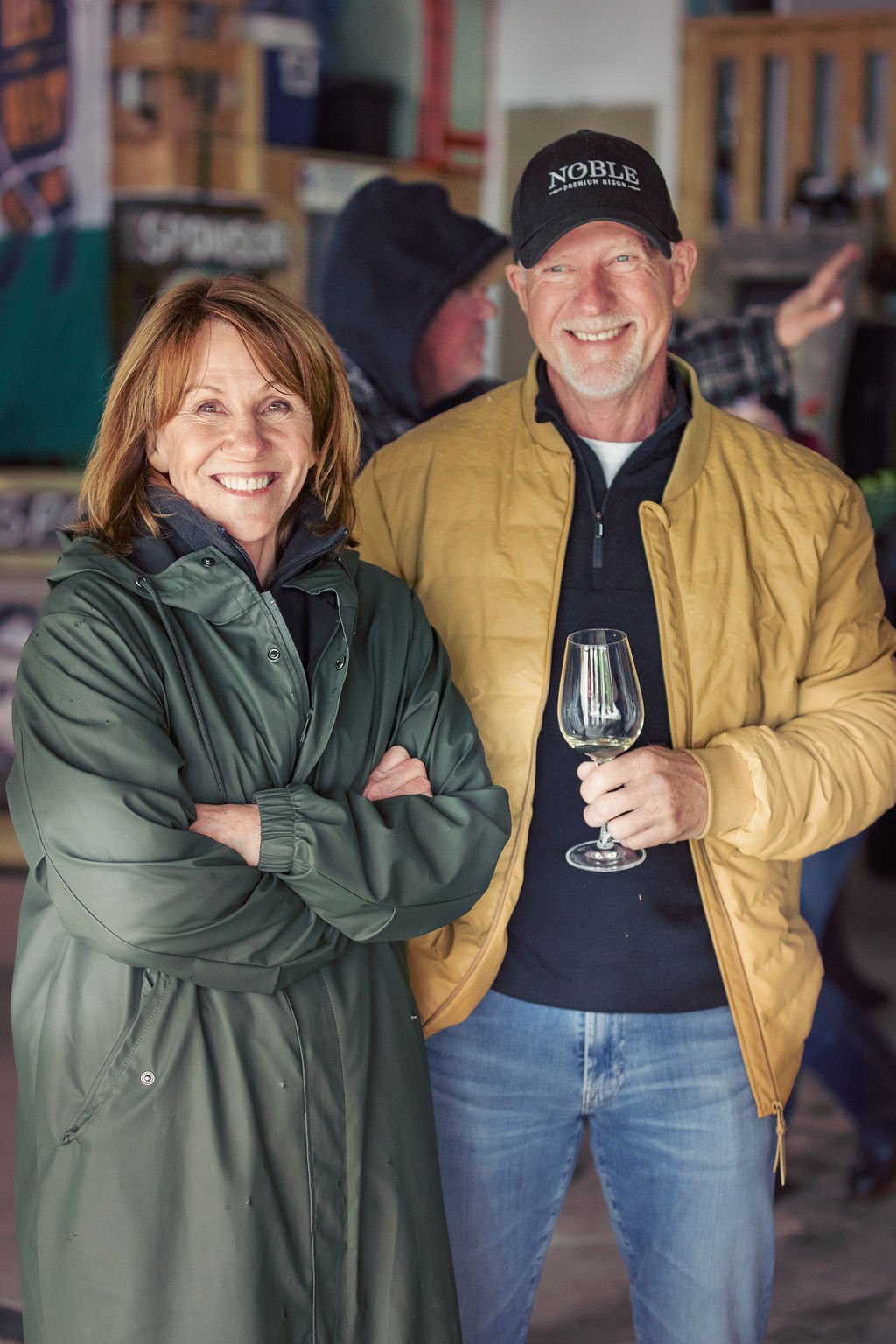 A smiling woman with shoulder-length brown hair wearing a green raincoat and a man with a gray beard wearing a yellow jacket and a black cap holding a glass of white wine, standing indoors.