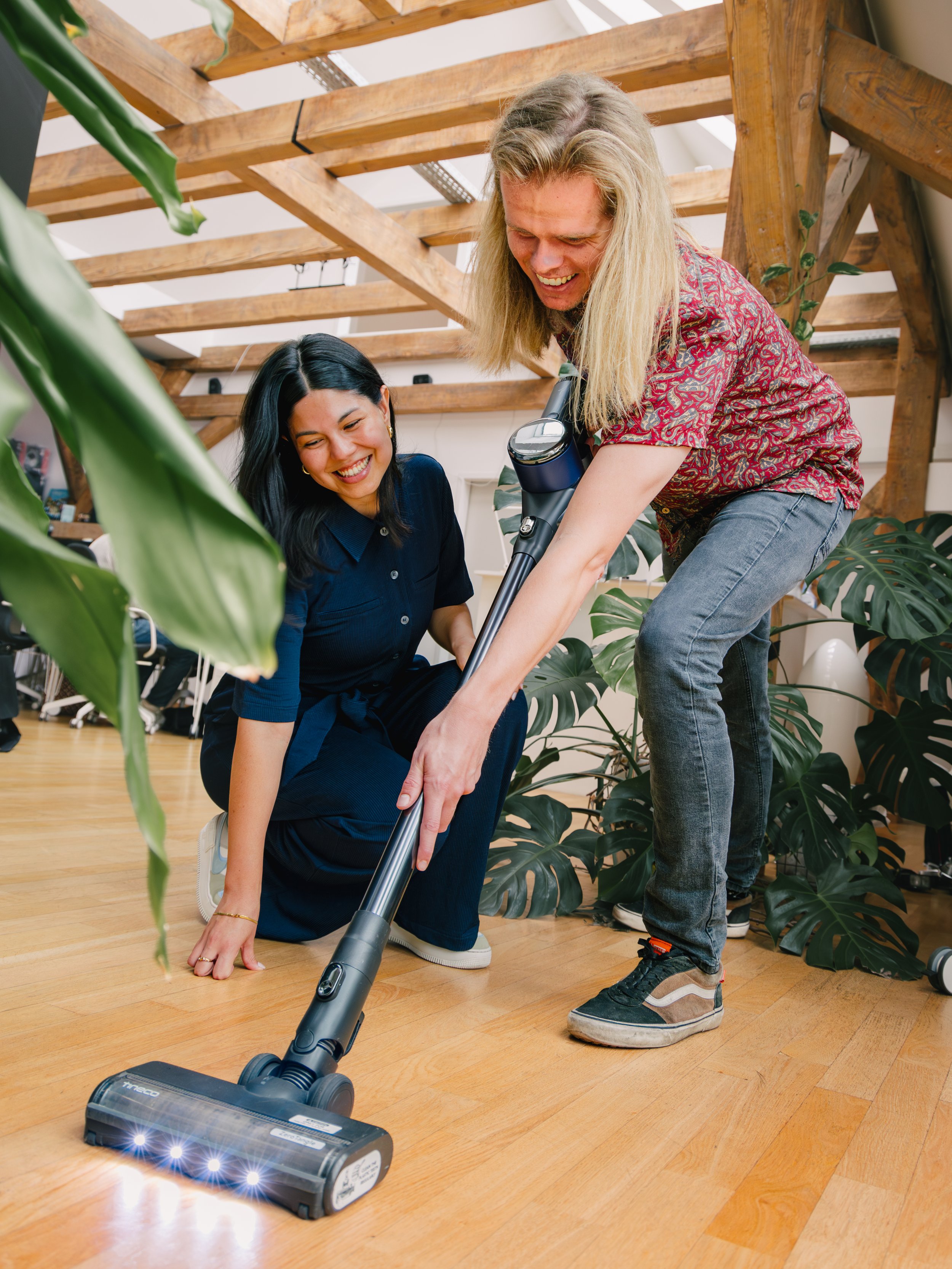 Two people are cleaning a hardwood floor with a vacuum cleaner inside a room with wooden beams and large green leafy plants. One person is kneeling on the ground, while the other is standing and leaning forward to vacuum.