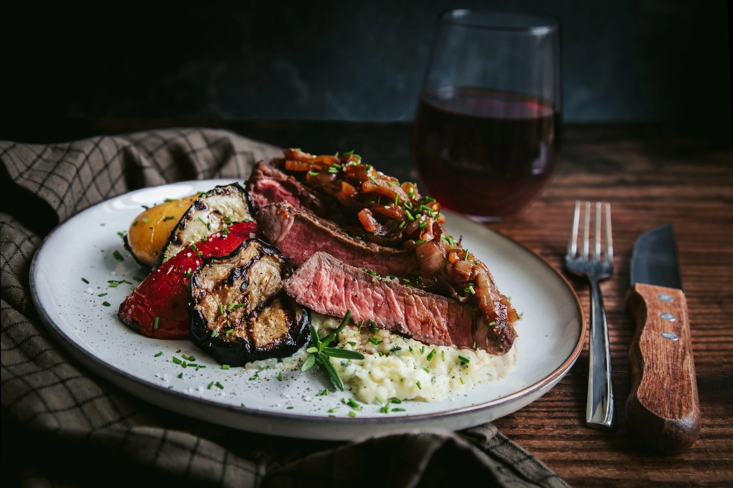 A plate of cooked steak, grilled eggplant, roasted red pepper, and mashed potatoes garnished with chopped chives, with a glass of red wine in the background on a wooden table.
