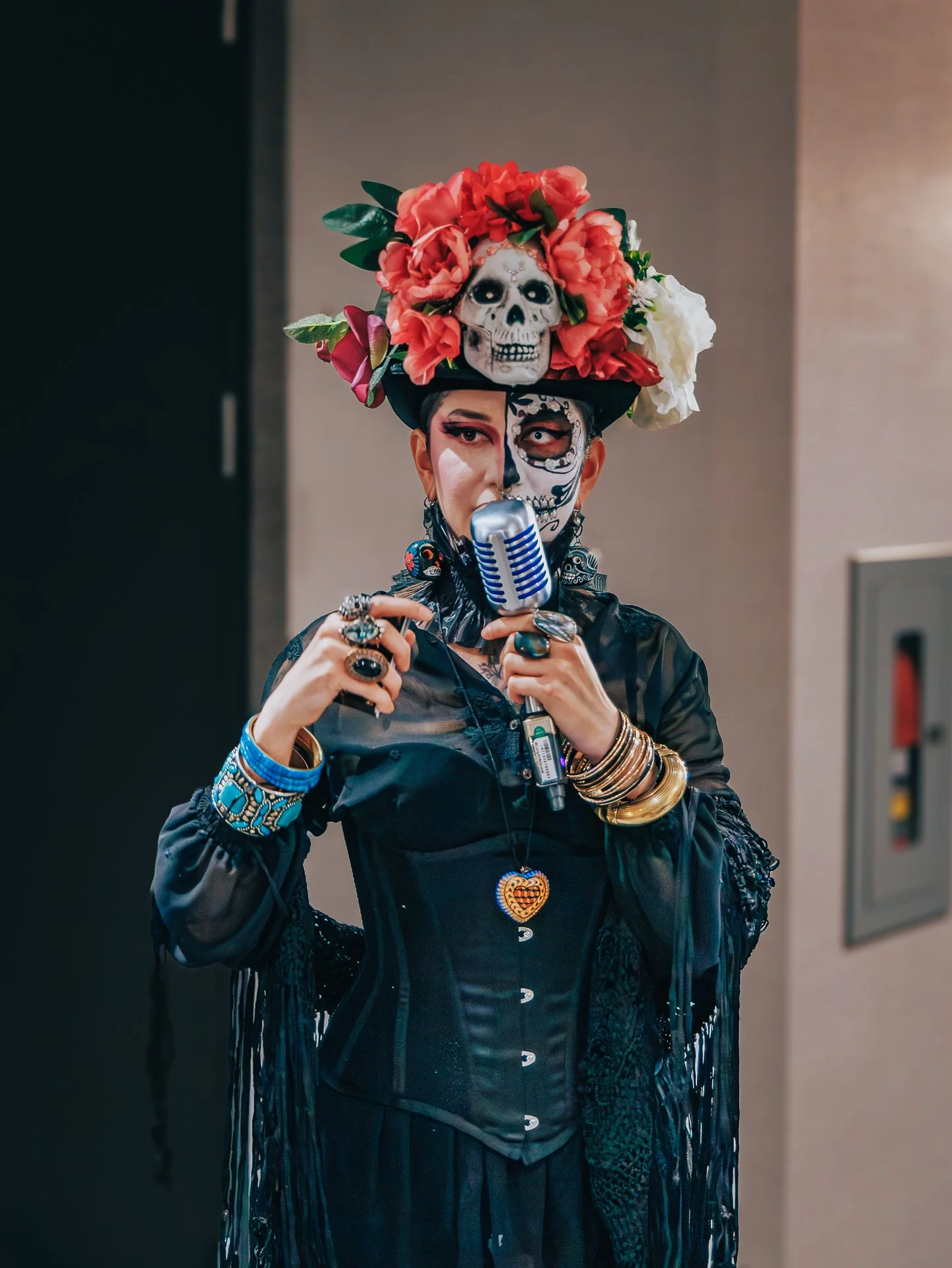 Person wearing Day of the Dead makeup and a large floral hat with skull and flower decorations, holding a vintage microphone.