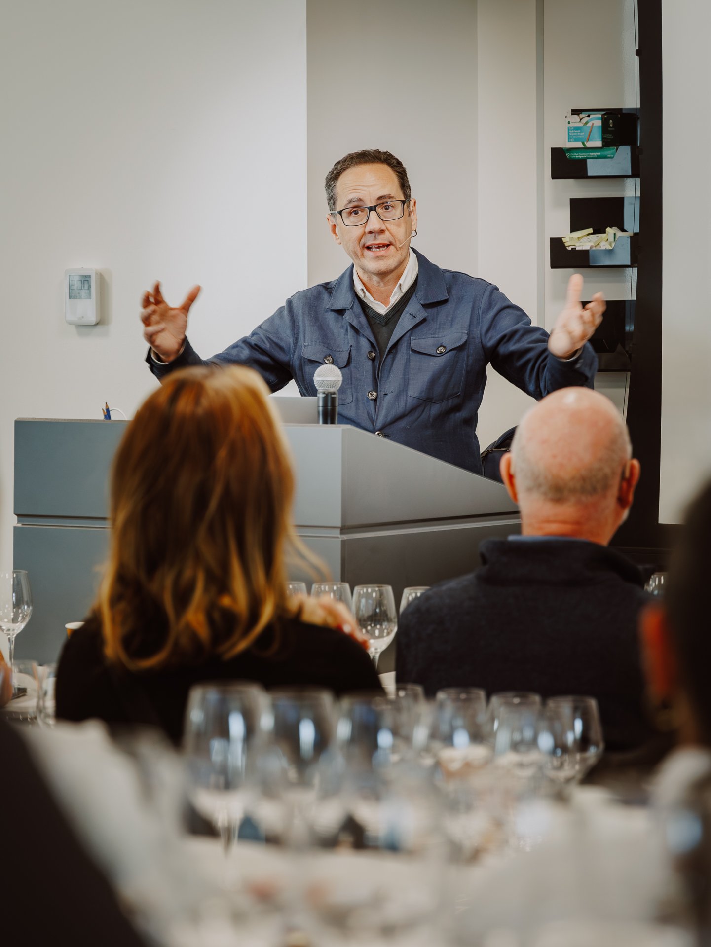 A man giving a presentation at a conference, standing behind a lectern with a microphone, gesturing with his hands, audience members seated in front of him, with wine glasses on the table.