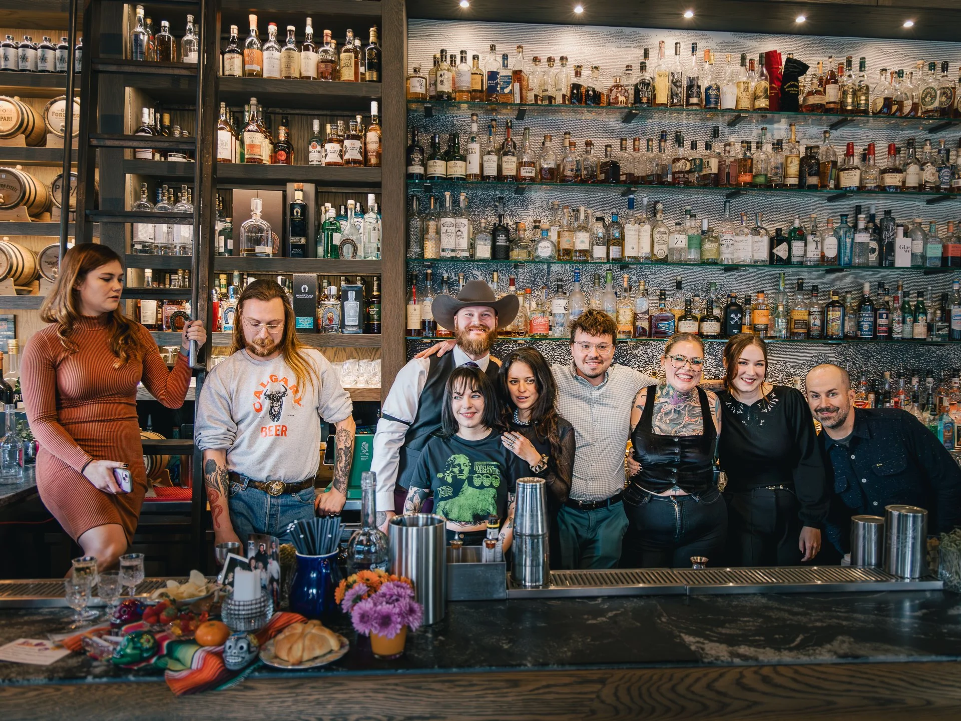 Group of people gathered at a bar with a large collection of liquor bottles behind them. There are smiling and serious expressions, with some people posing for the photo and others looking elsewhere. The bar features a marble counter with colorful fl