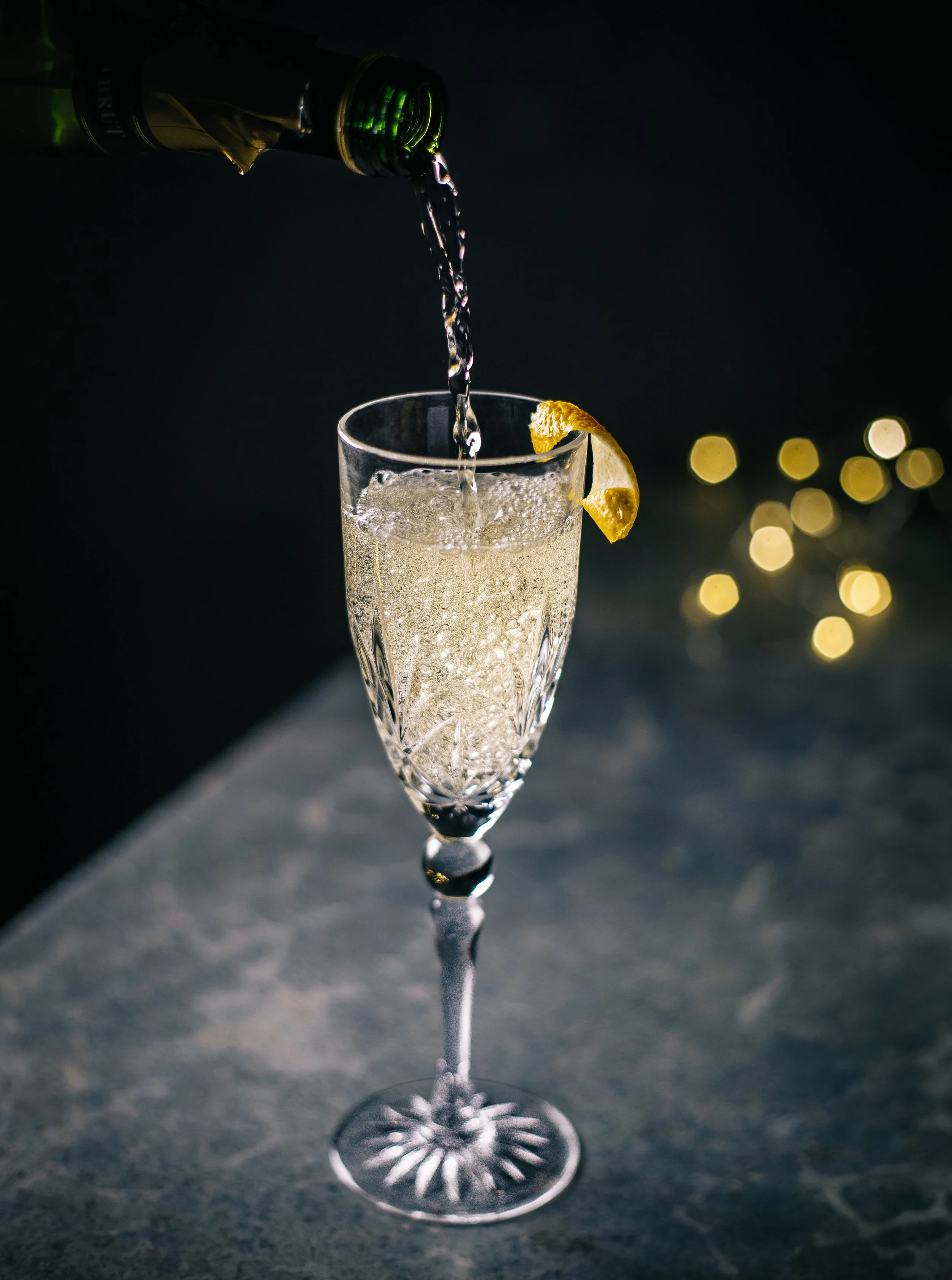 A glass of champagne with a lemon wedge on the rim, being poured from a bottle in a dark setting with bokeh lights in the background.