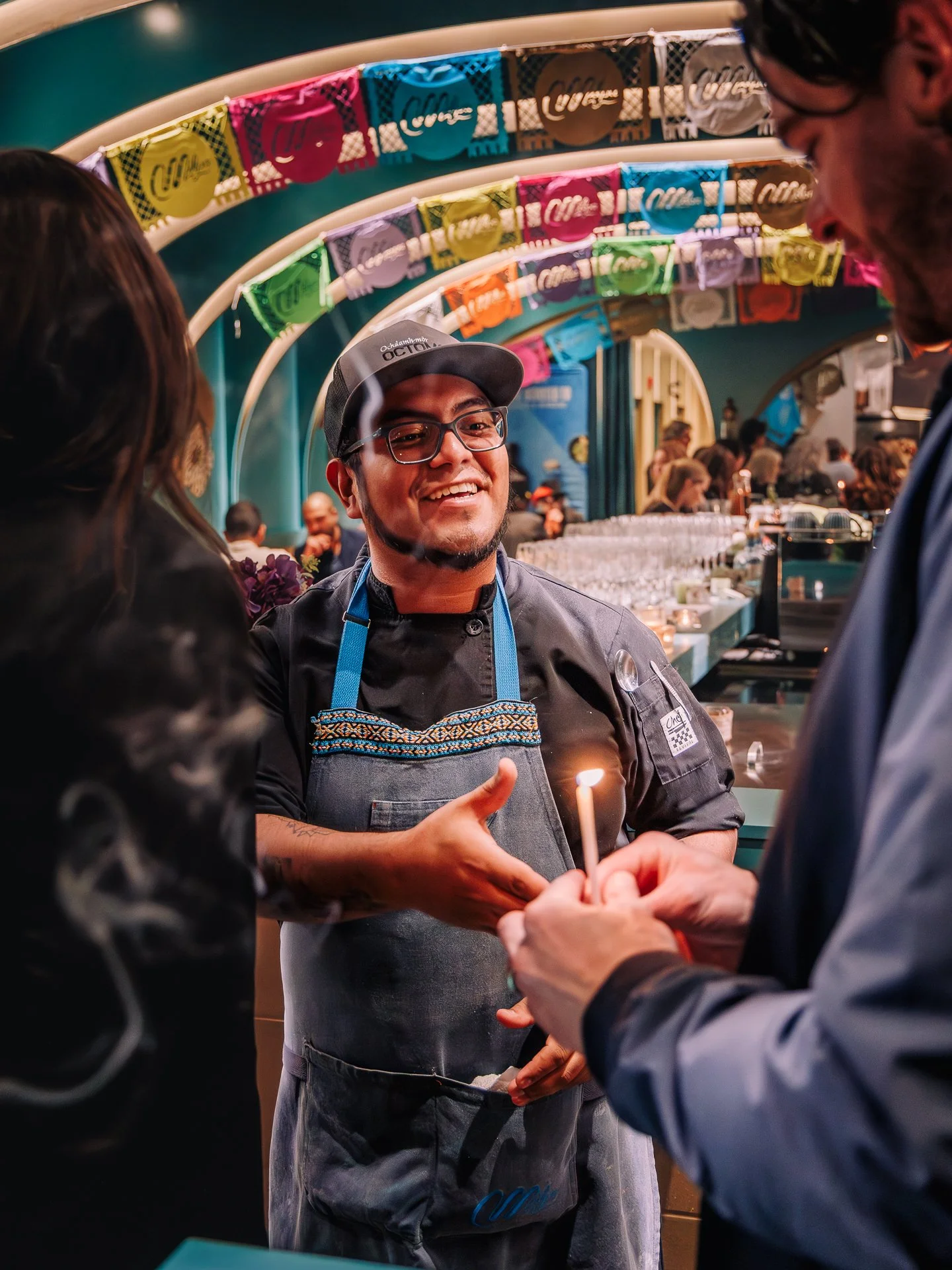 A man with glasses and a cap is smiling and shaking hands with another person at an event. The background has colorful banners and a busy atmosphere.