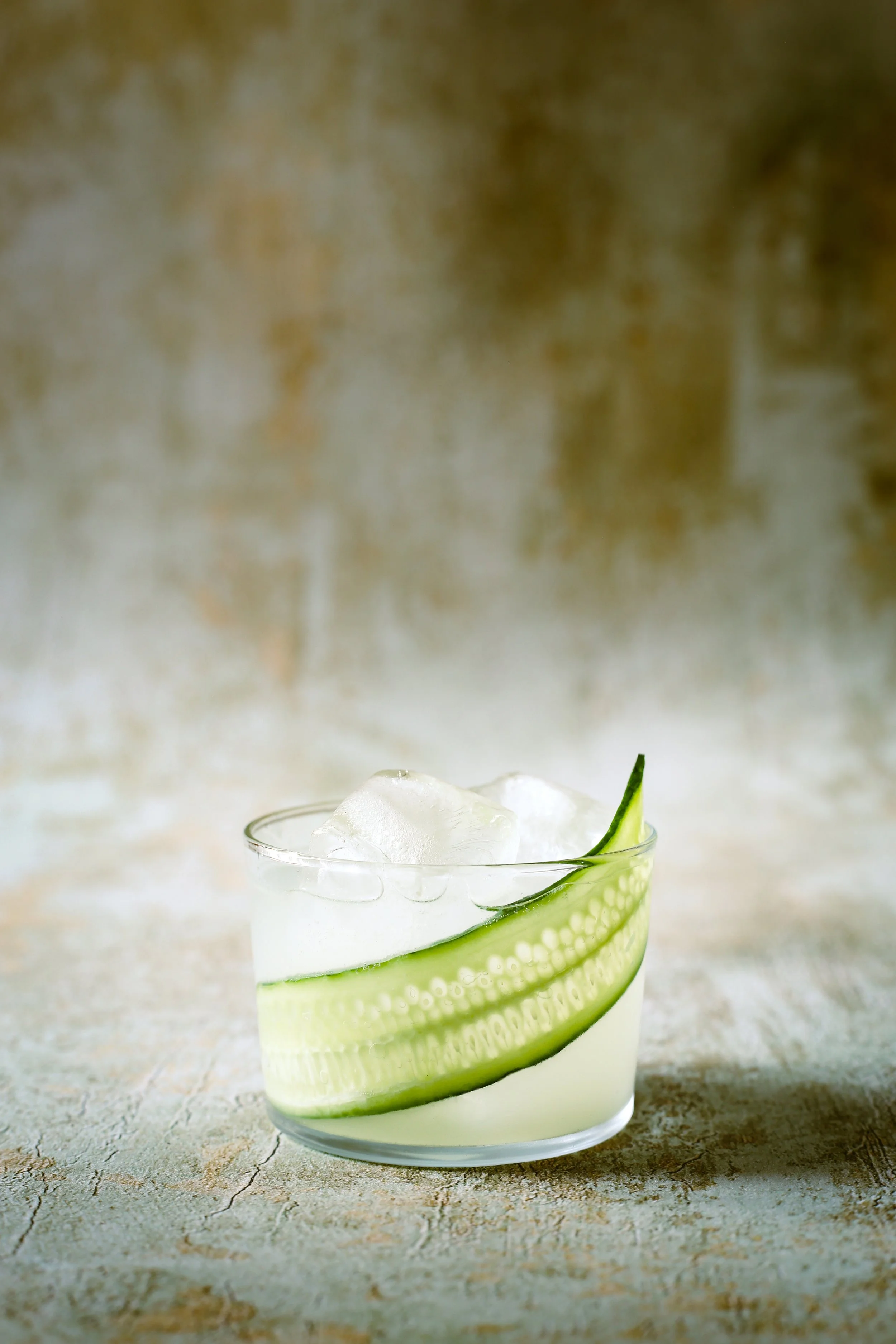 A glass of cucumber-infused beverage with ice cubes and a cucumber slice garnish placed on a textured surface.