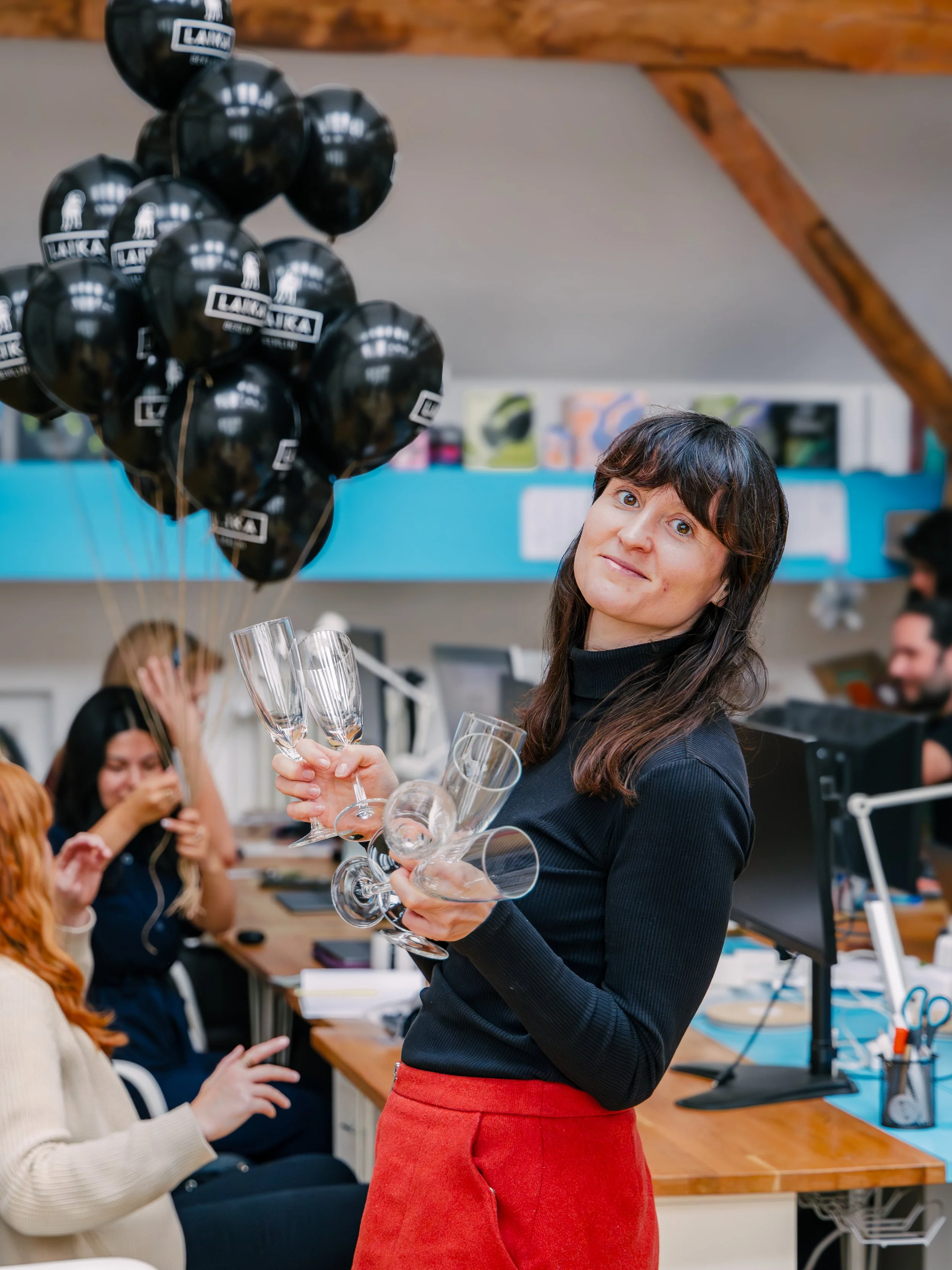 A woman in a black top and red pants holding champagne glasses in a room with people in the background, some holding balloons and others taking photos.