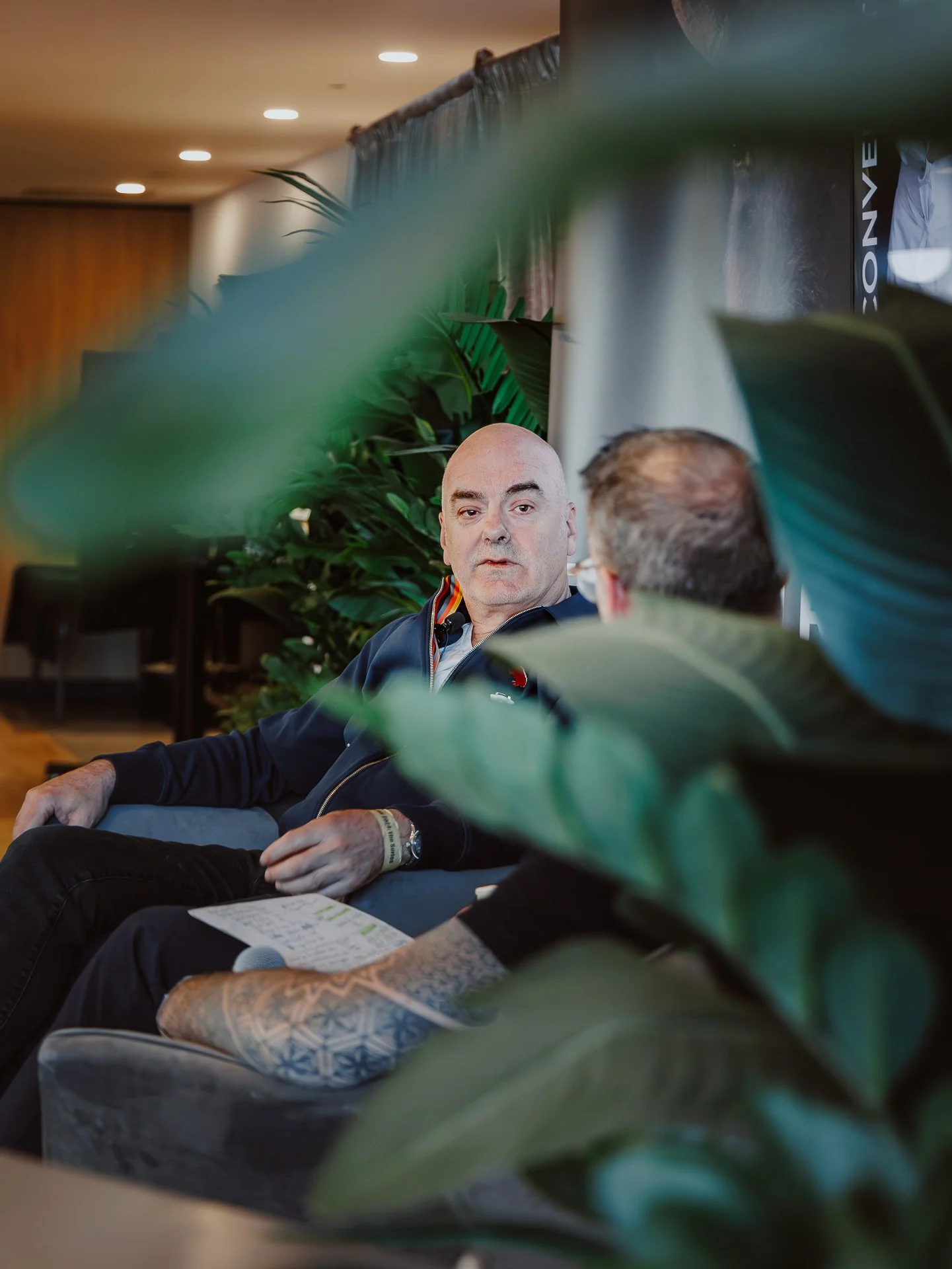Two men sitting in a room, partially obscured by green large leafy plants, engaged in conversation. One man has a shaved head and is wearing a dark jacket, while the other has short hair and glasses. The setting appears to be indoors, with a banner a