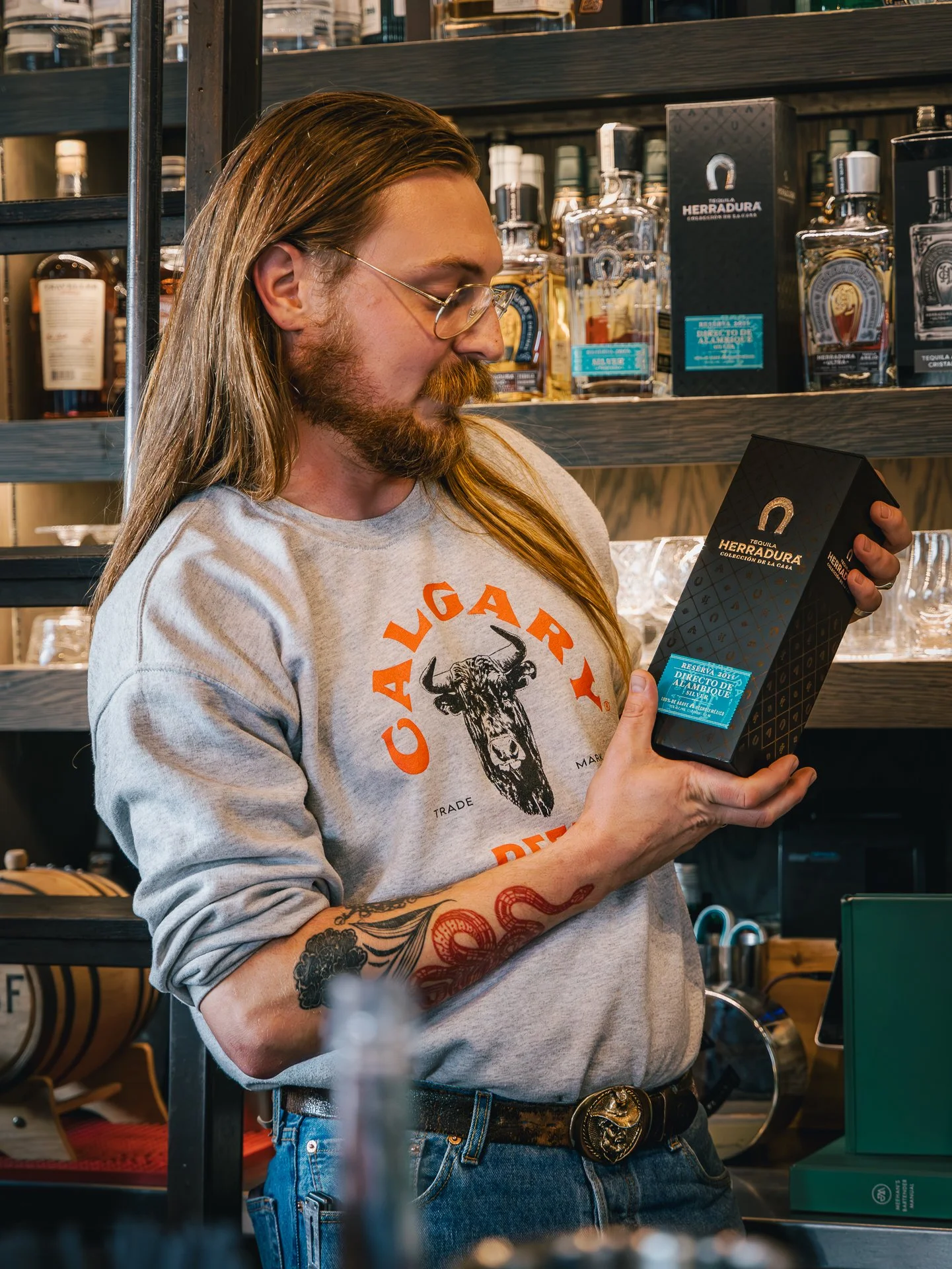 A man with long hair, glasses, and a beard holding a black box labeled 'HERRADURA' while standing in front of shelves with bottles of liquor.