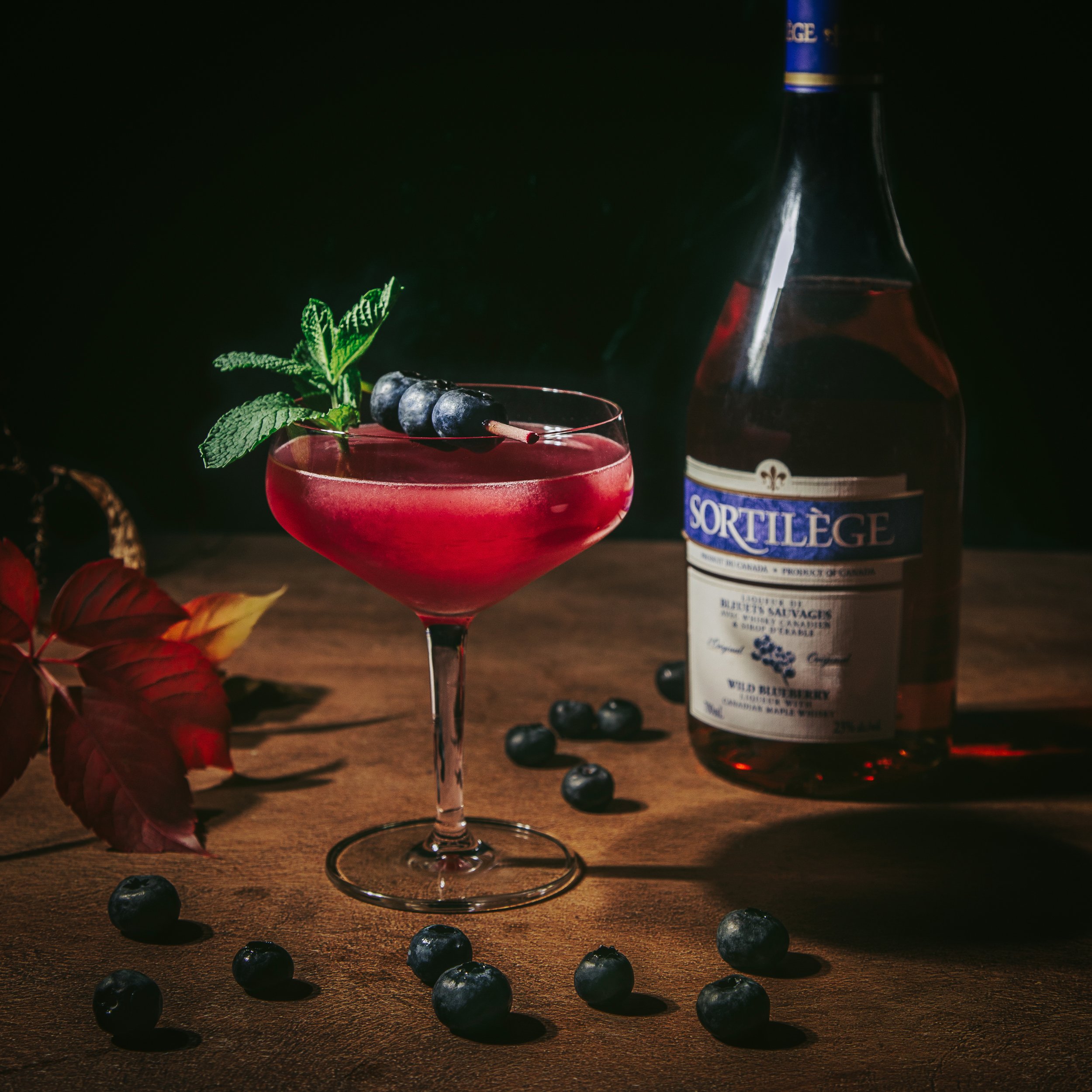 A glass of red beverage garnished with mint leaves and blueberries, with a bottle of alcohol in the background, placed on a wooden surface with scattered blueberries.