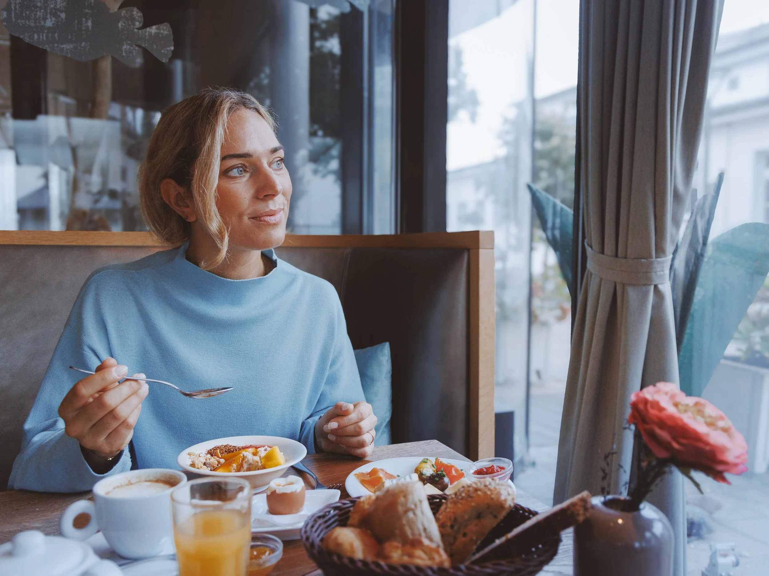 A woman with blonde hair and blue eyes sitting at a table with breakfast foods, holding a spoon. The table has various dishes including pastries, fruit, a cup of coffee, and orange juice. There is a window with curtains behind her, and outside, trees