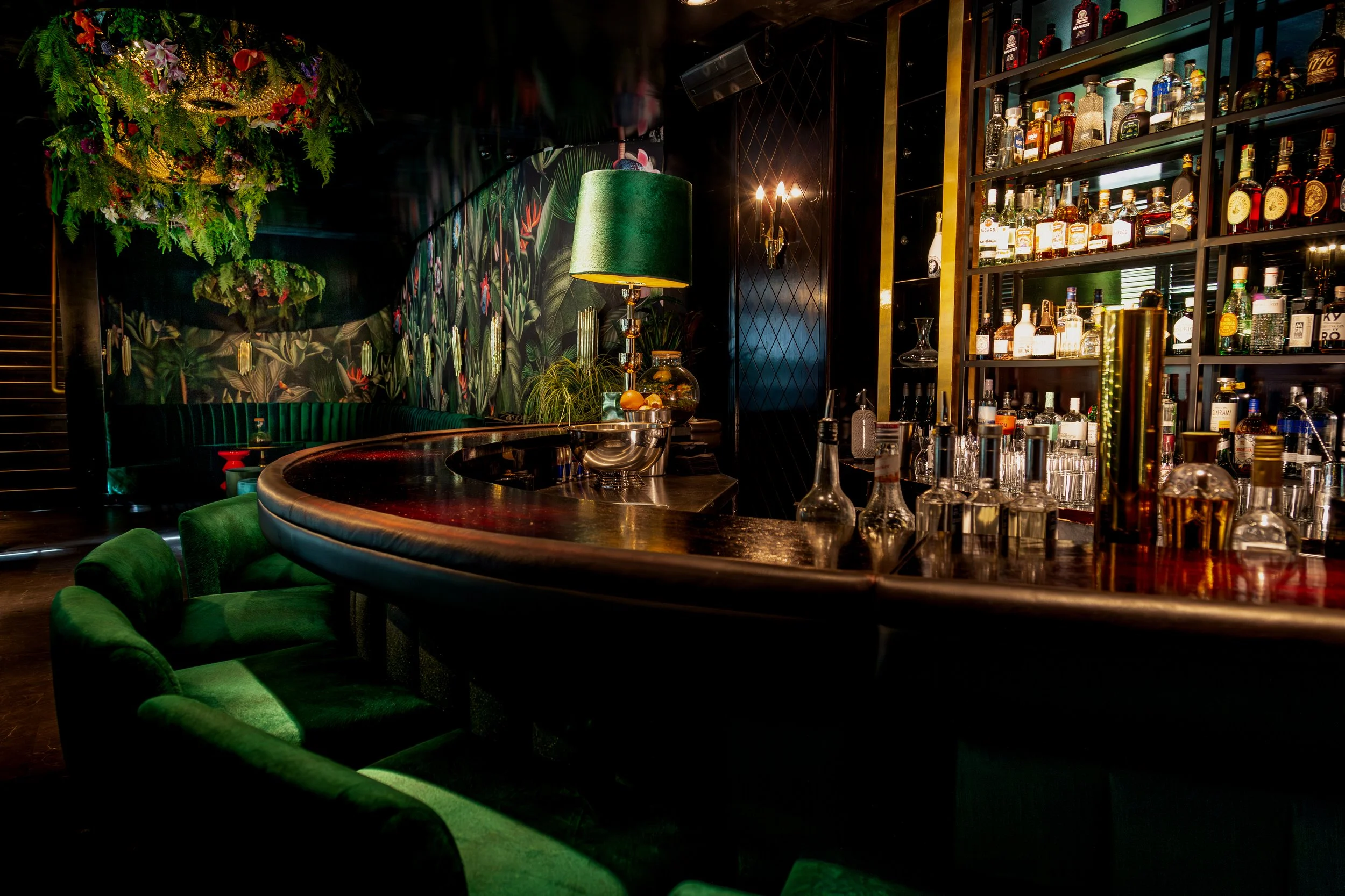 Interior of a stylish bar with green velvet chairs around a curved dark counter, lush tropical wallpaper, a gaze green lamp, and shelves of liquor bottles with warm lighting.