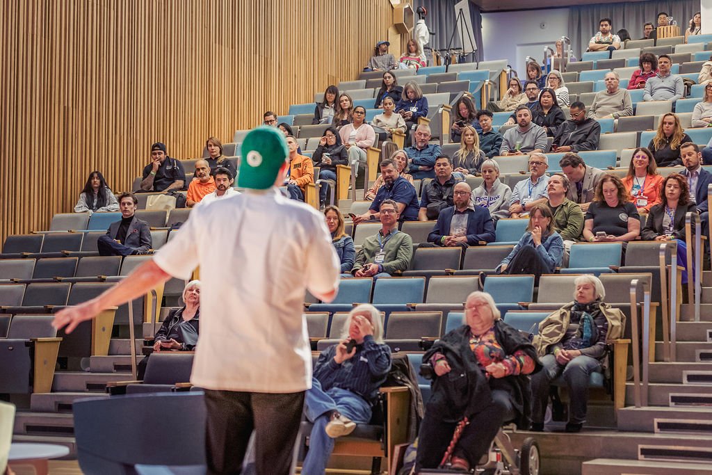 A speaker in a medical coat and green cap stands in front of an audience in a lecture hall, with attendees seated in tiered rows.
