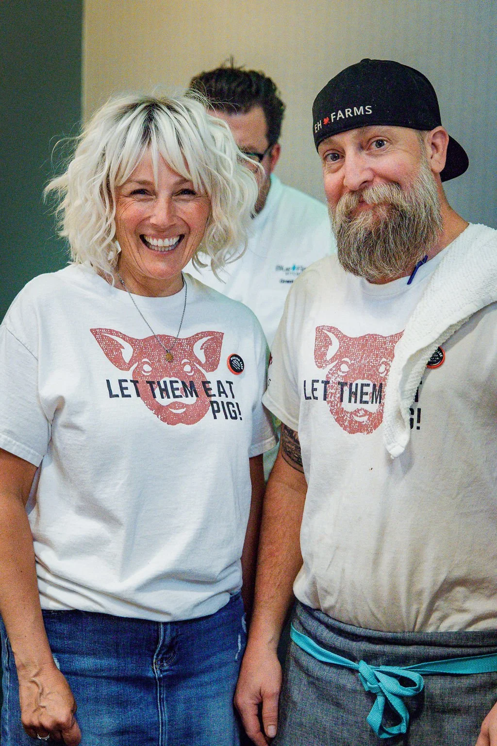 Two smiling people, a woman with blonde curly hair and a man with a beard and black cap, wearing white T-shirts with a pig graphic and the phrase "Let Them Eat Pig!"