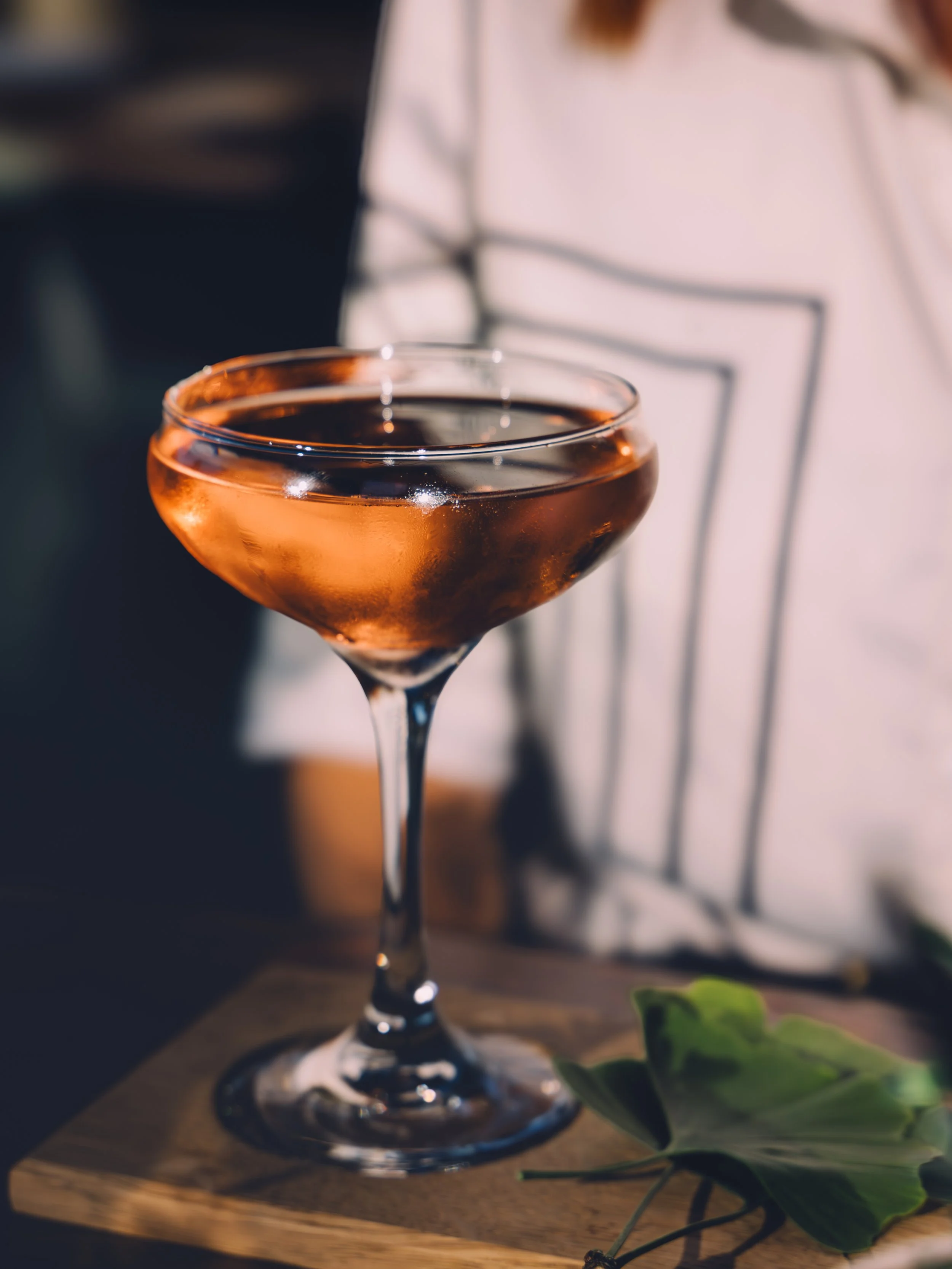 A close-up of a pink champagne coupe glass filled with a pink beverage, placed on a wooden surface with some green leaves nearby.