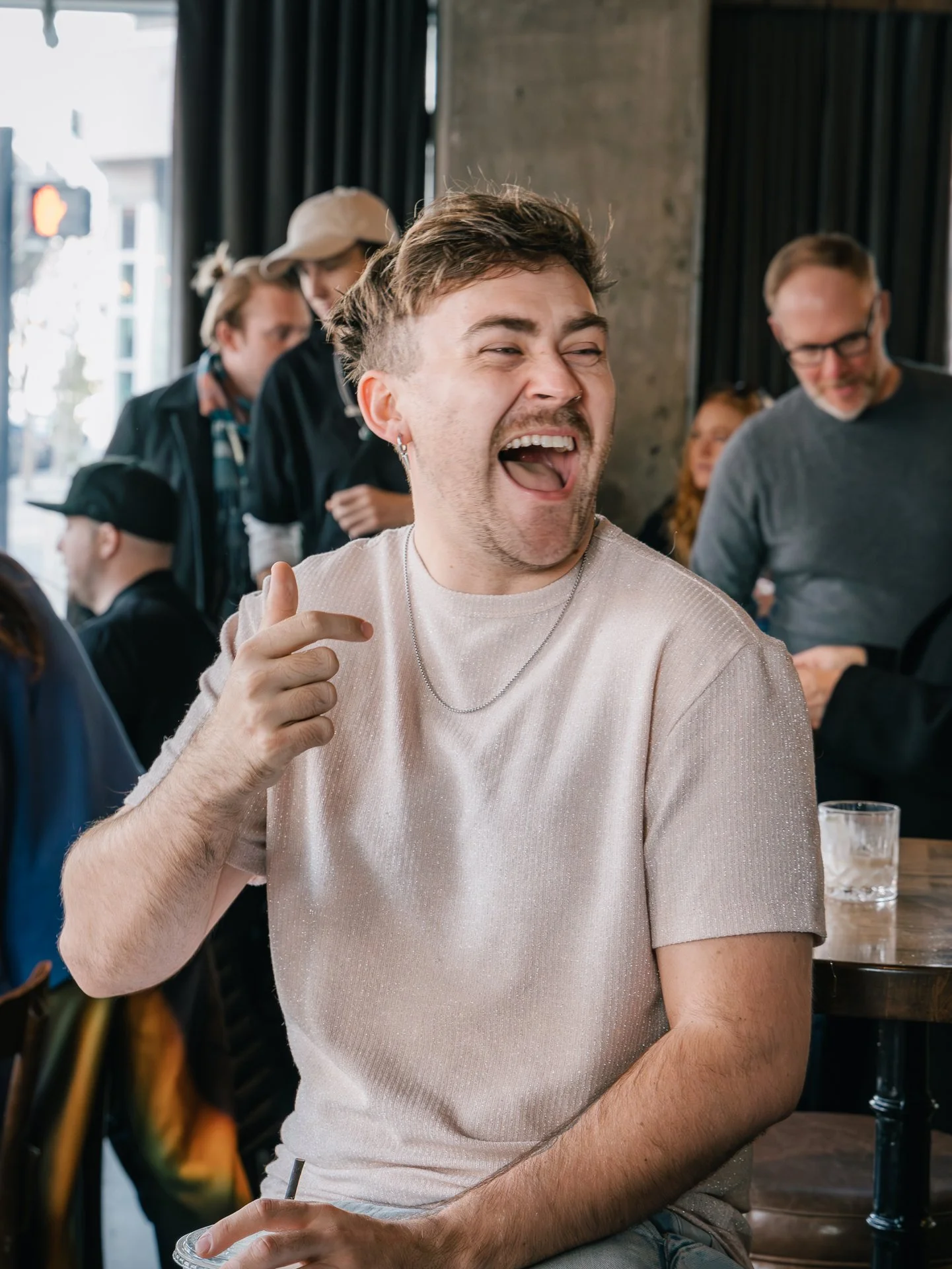 A young man with short brown hair and earrings laughing and pointing with his index finger, sitting in a lively indoor setting with other people in the background.
