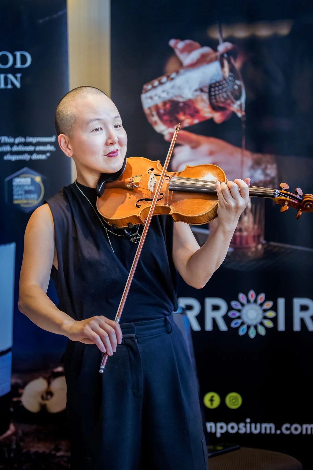 A woman with a shaved head playing a violin in front of a promotional banner.