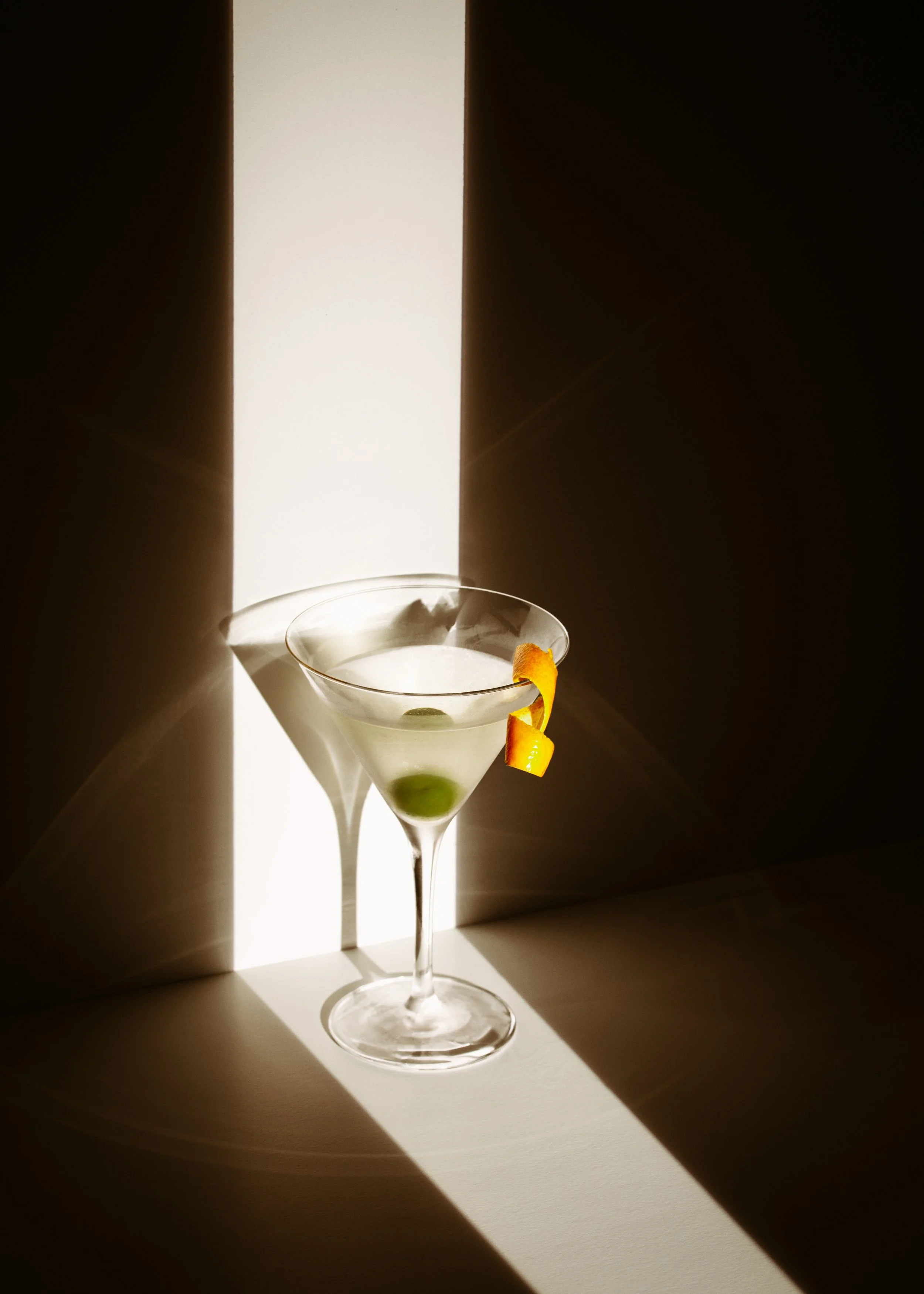 A martini glass with green olives and an orange garnish, illuminated by dramatic light and shadow, creating an artistic effect.